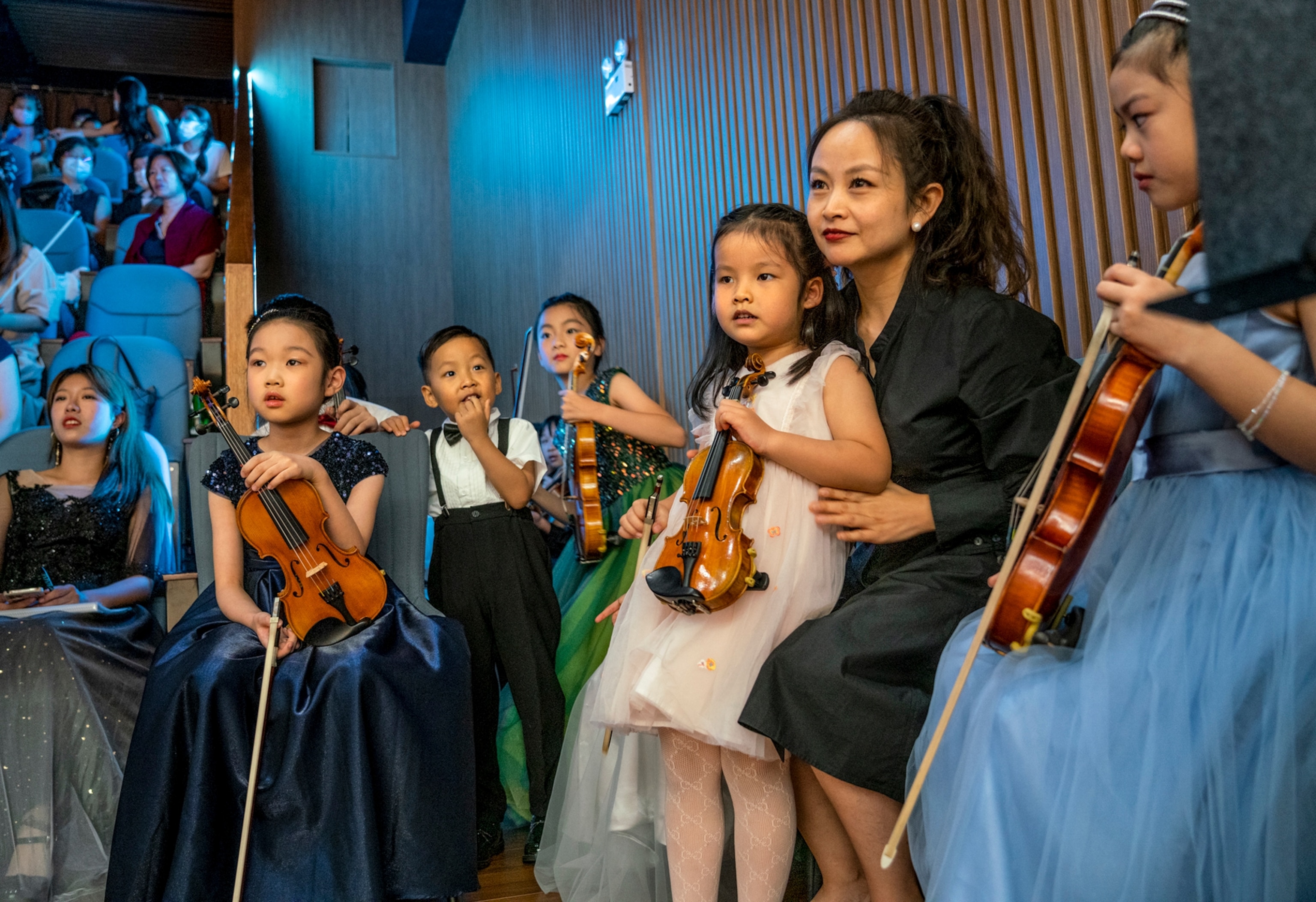 Picture of a group of children all with violins and dressed nicely sitting to the side of a stage.