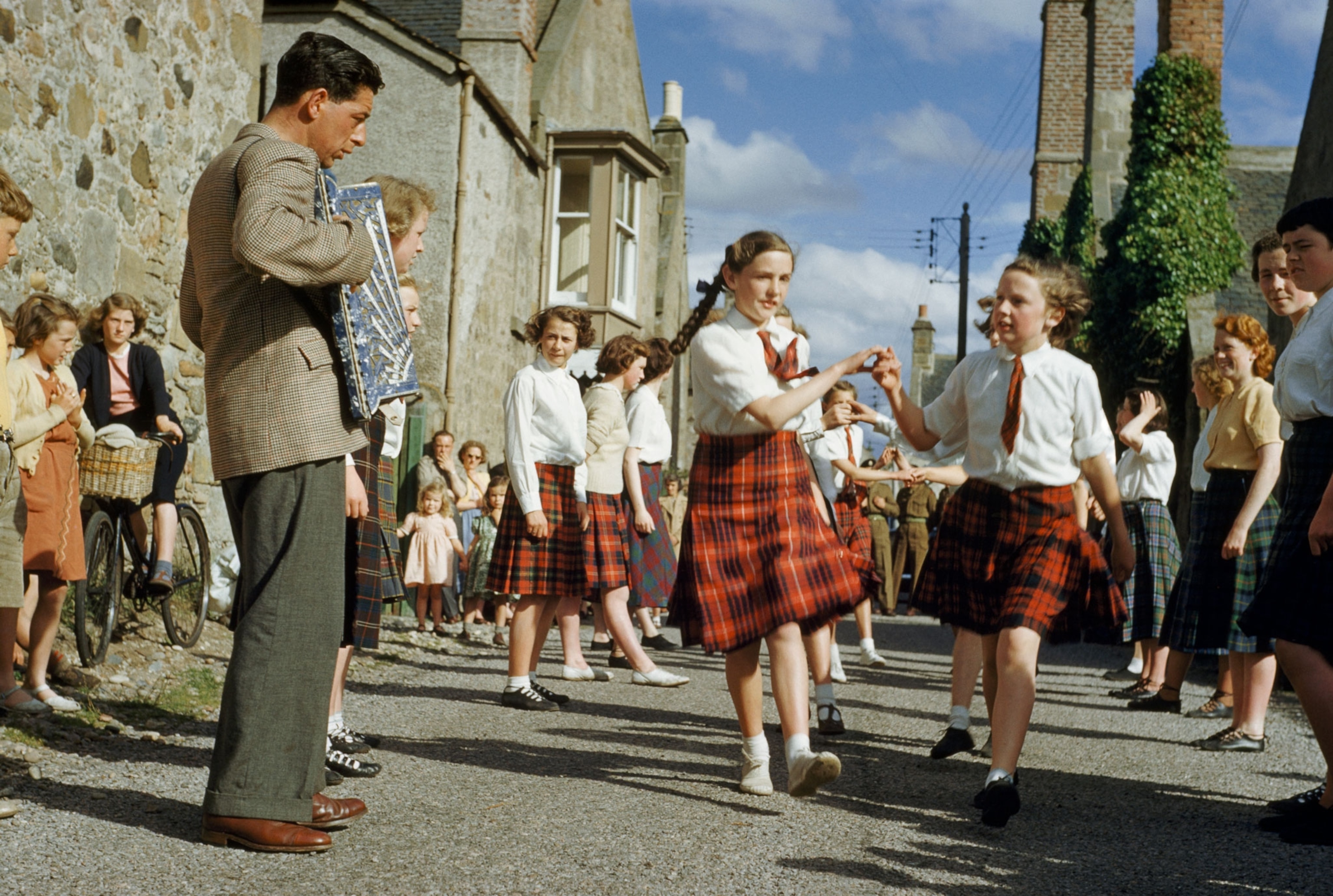 An accordionist plays for dancing schoolgirls in tartan plaid skirts.