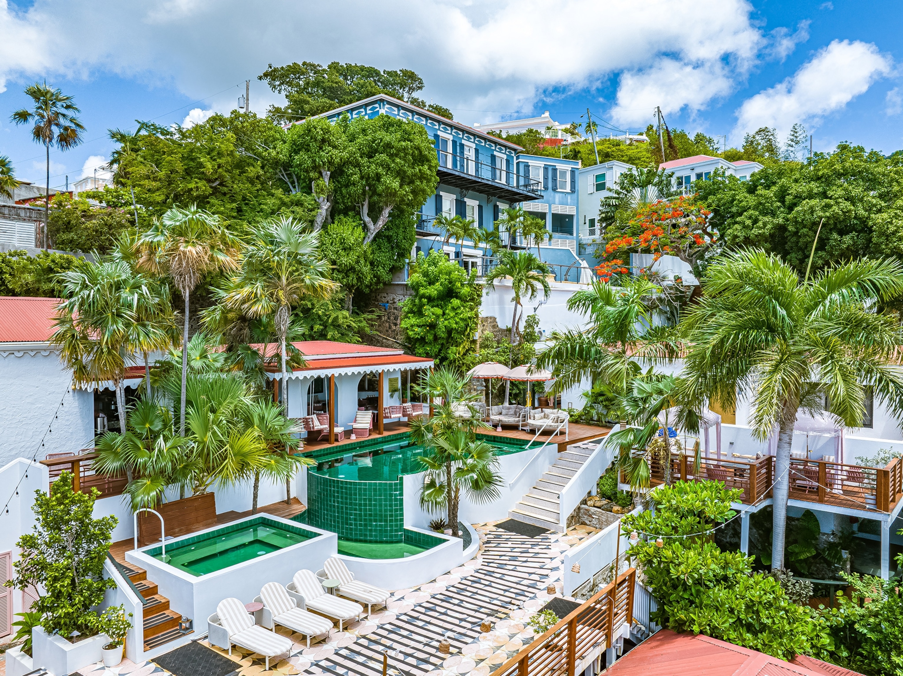 A view of the the Pink Palm Hotel, made up of several mid-20th-century buildings and filled with trees and a swimming pool in the center.