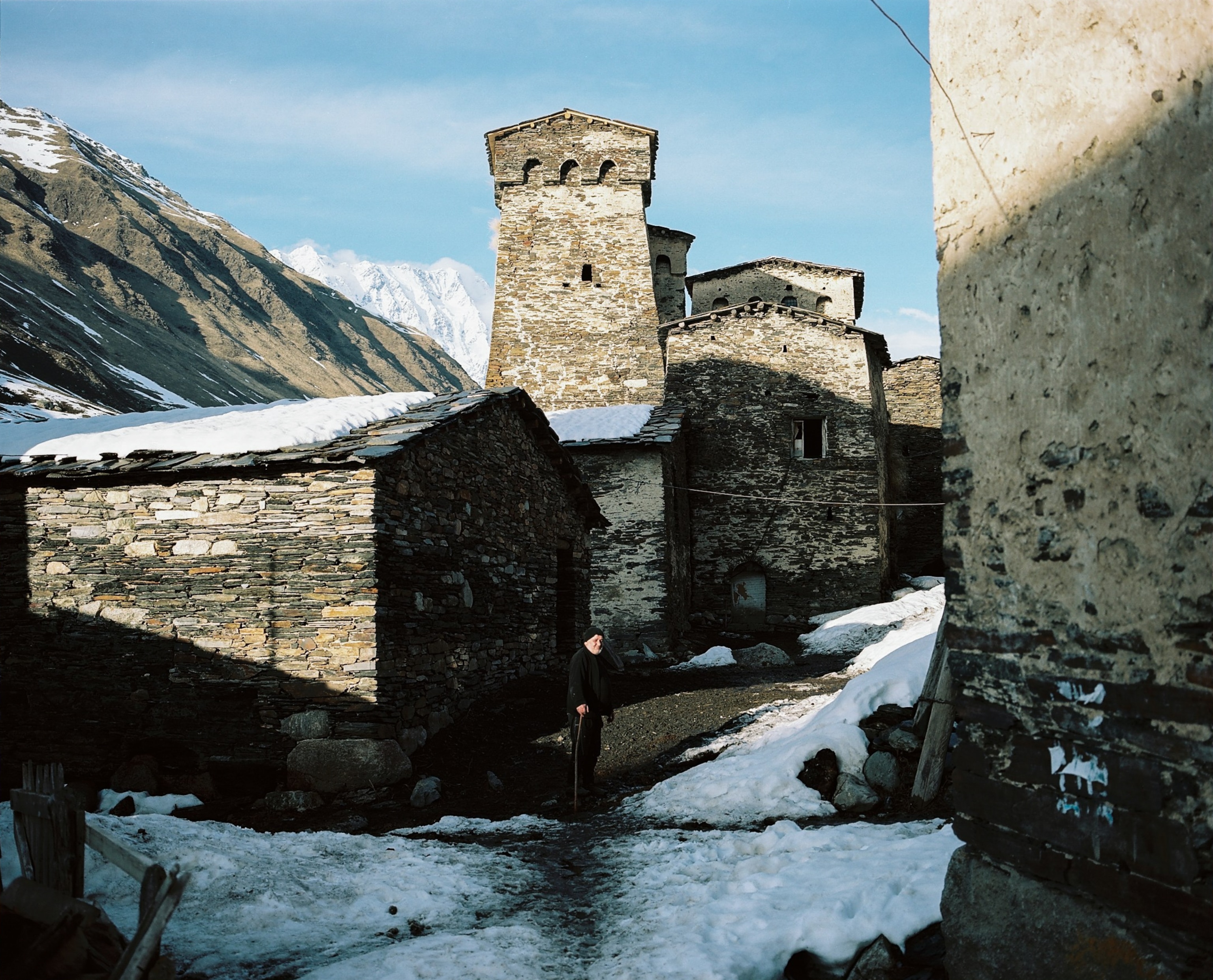 A man stands in a patch of sun among the towers of Ushguli