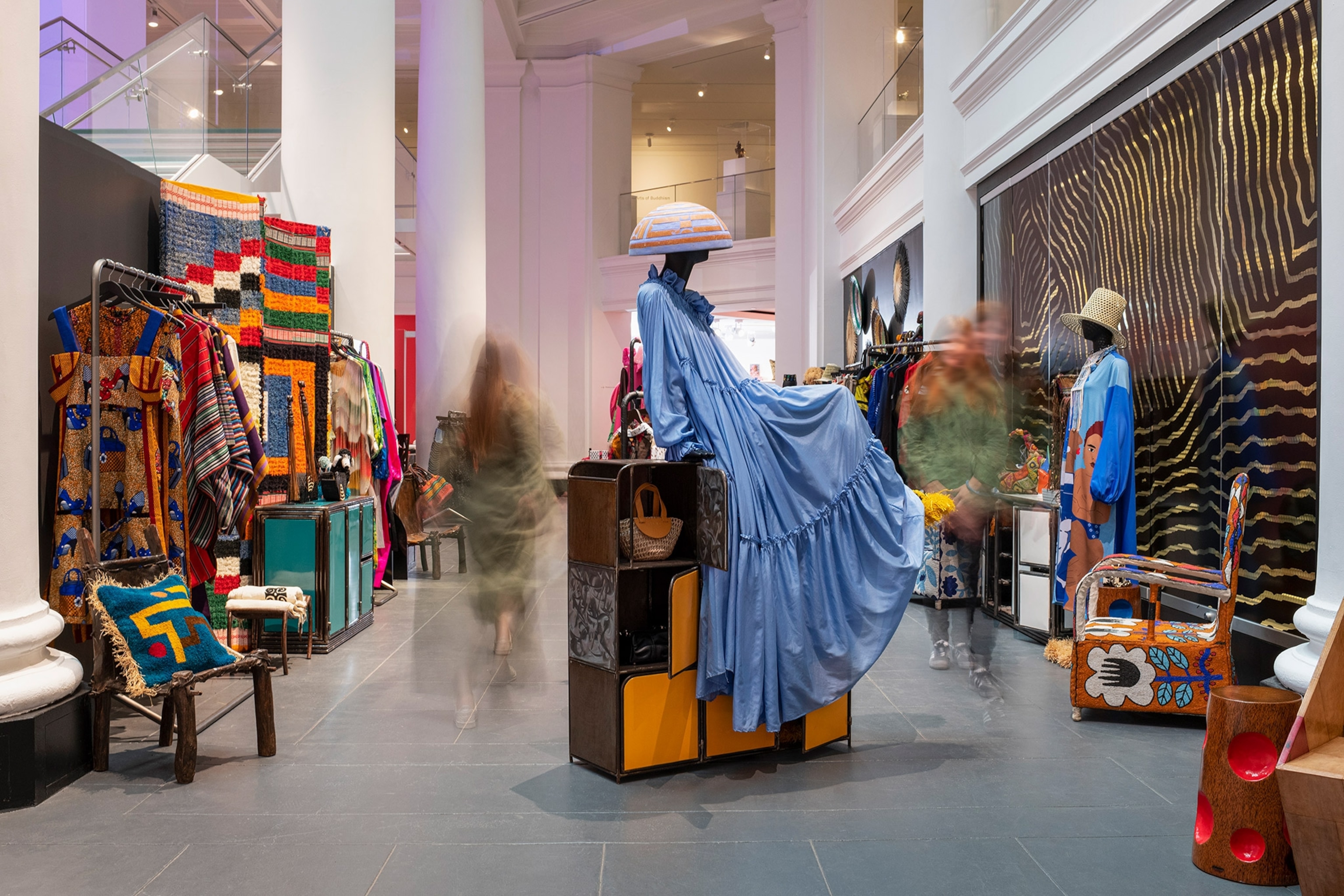 A manequin dons a blue dress and a hat, colorful patters are on the walls and various items int he exhibition.