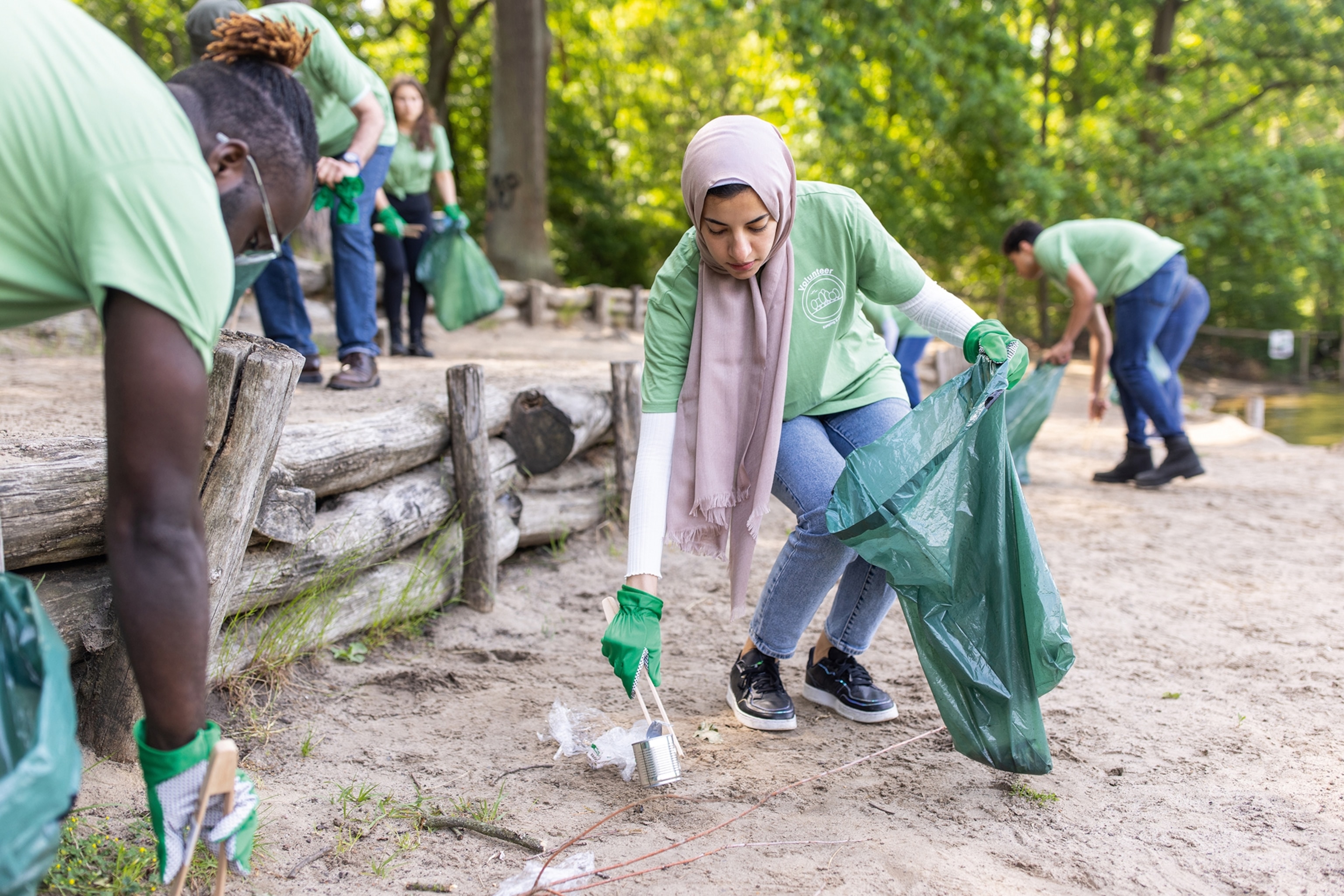 A female volunteer is picking up litter as part of a clean-up project. She is wearing a green tshirt and a purple head scarf.