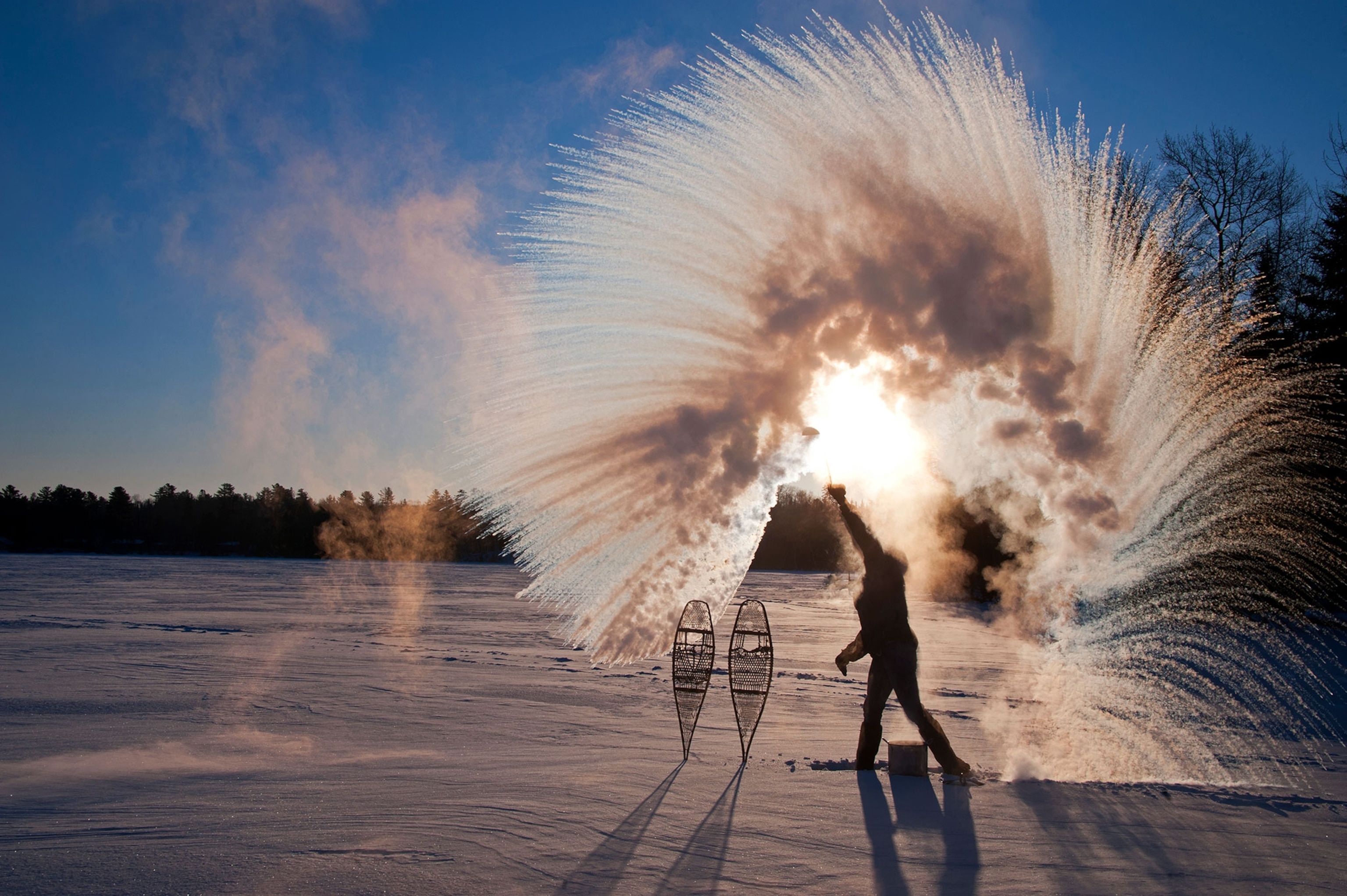 A winter scene in which a siluetted figure, standing in the middle of a snow covered field, tosses hot water, overhead, into the air to make a circular spray of frozen water. Two snow shoes are stuck in the snow nearby.