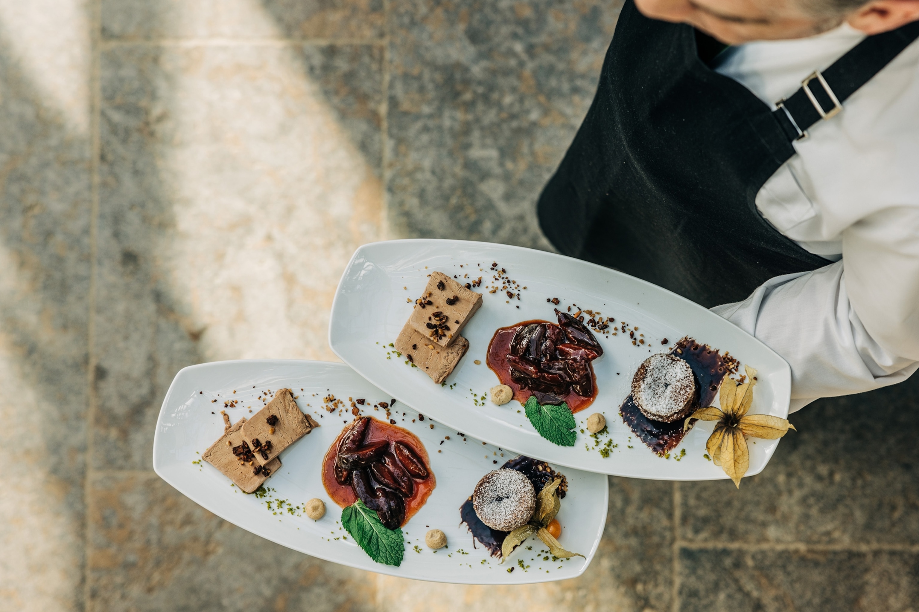 A waiter is holding two plates on one hand. They are a long rectangle shape, and have a selection of chocolate deserts on each.