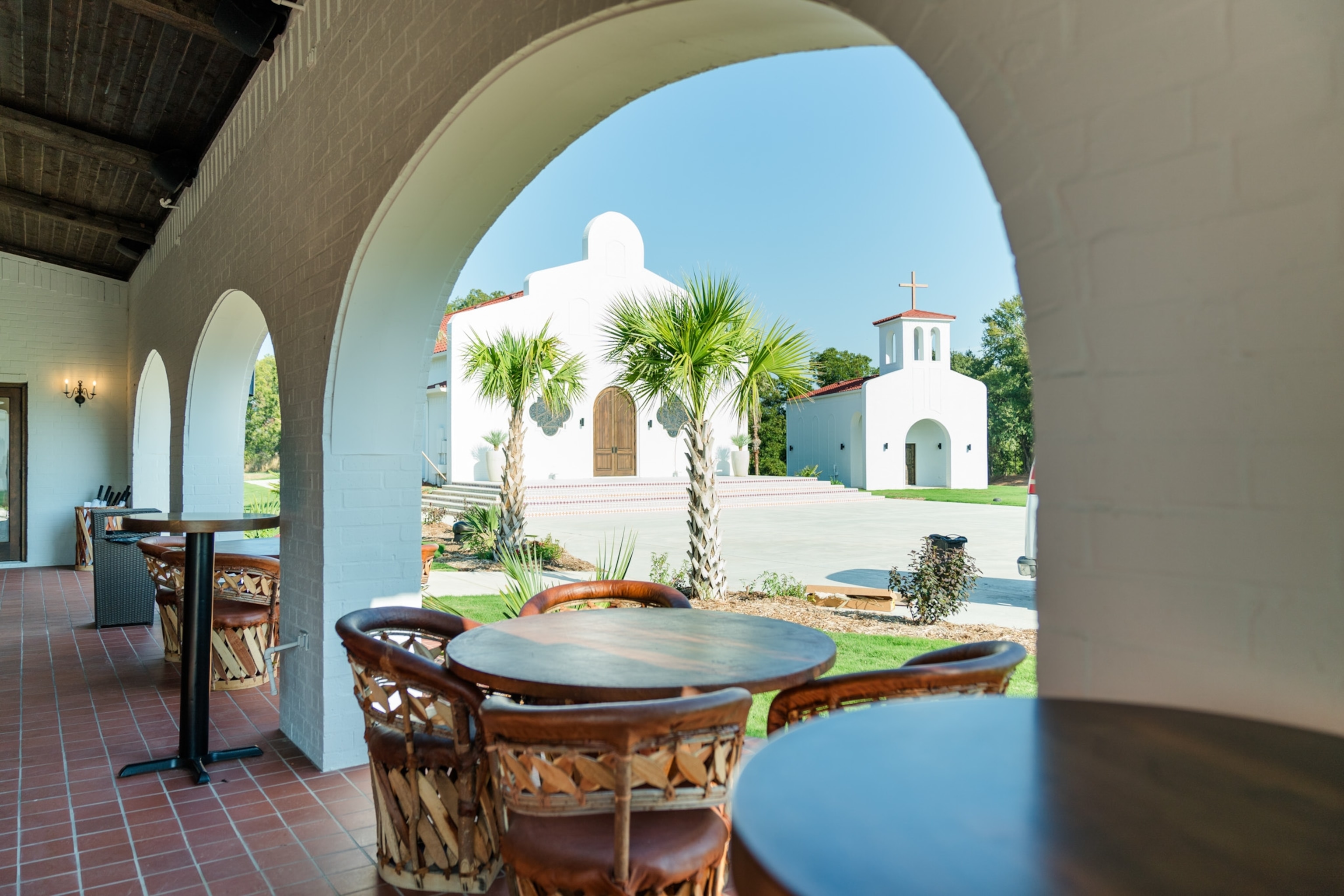 A patio with small tables and chairs with open archways looking through to two white chapels and palm trees.