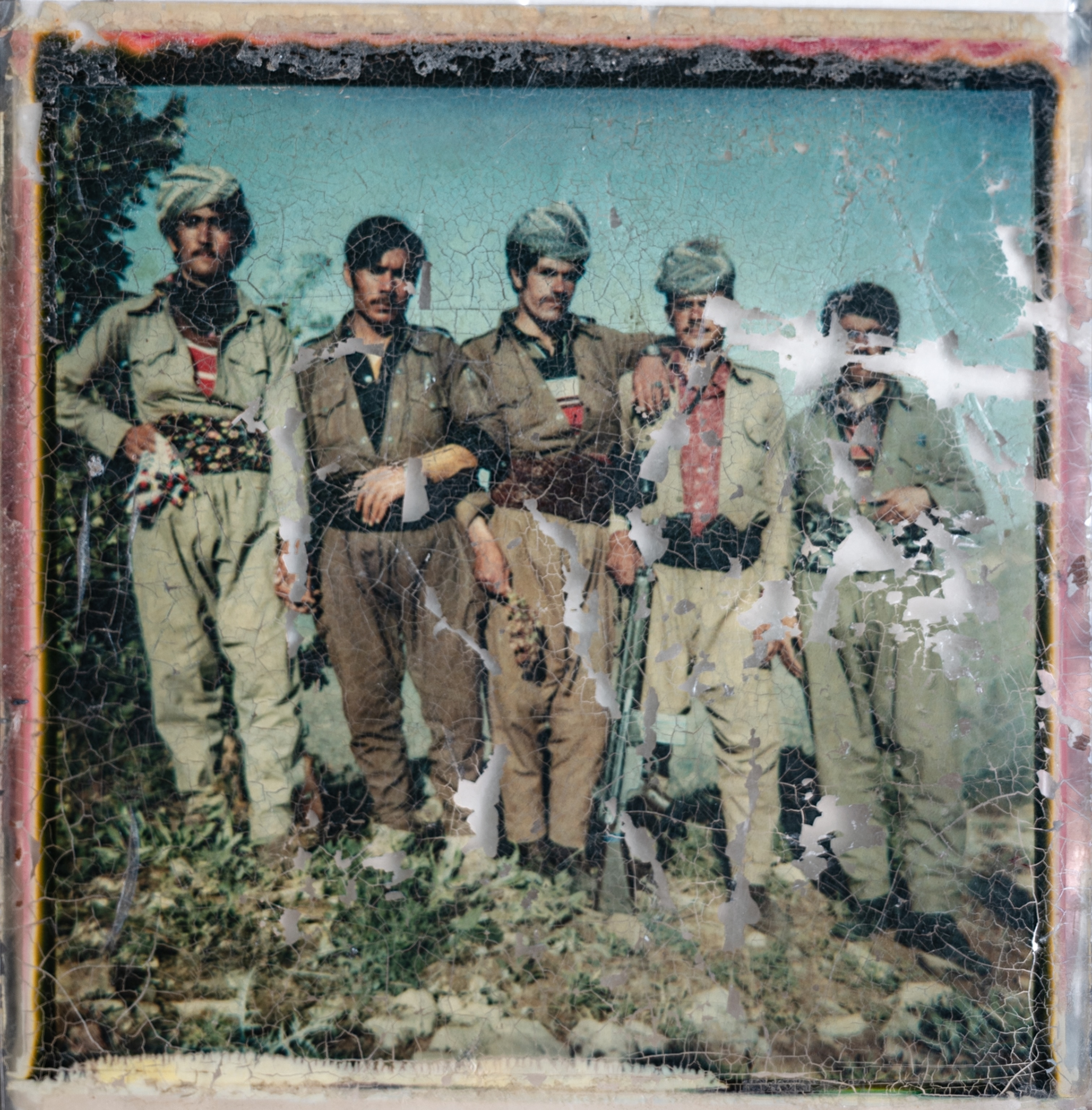 A nearly eroded square color photograph shows five soldiers in uniform gazing forward with a firearm at their waist in front of a blue sky.