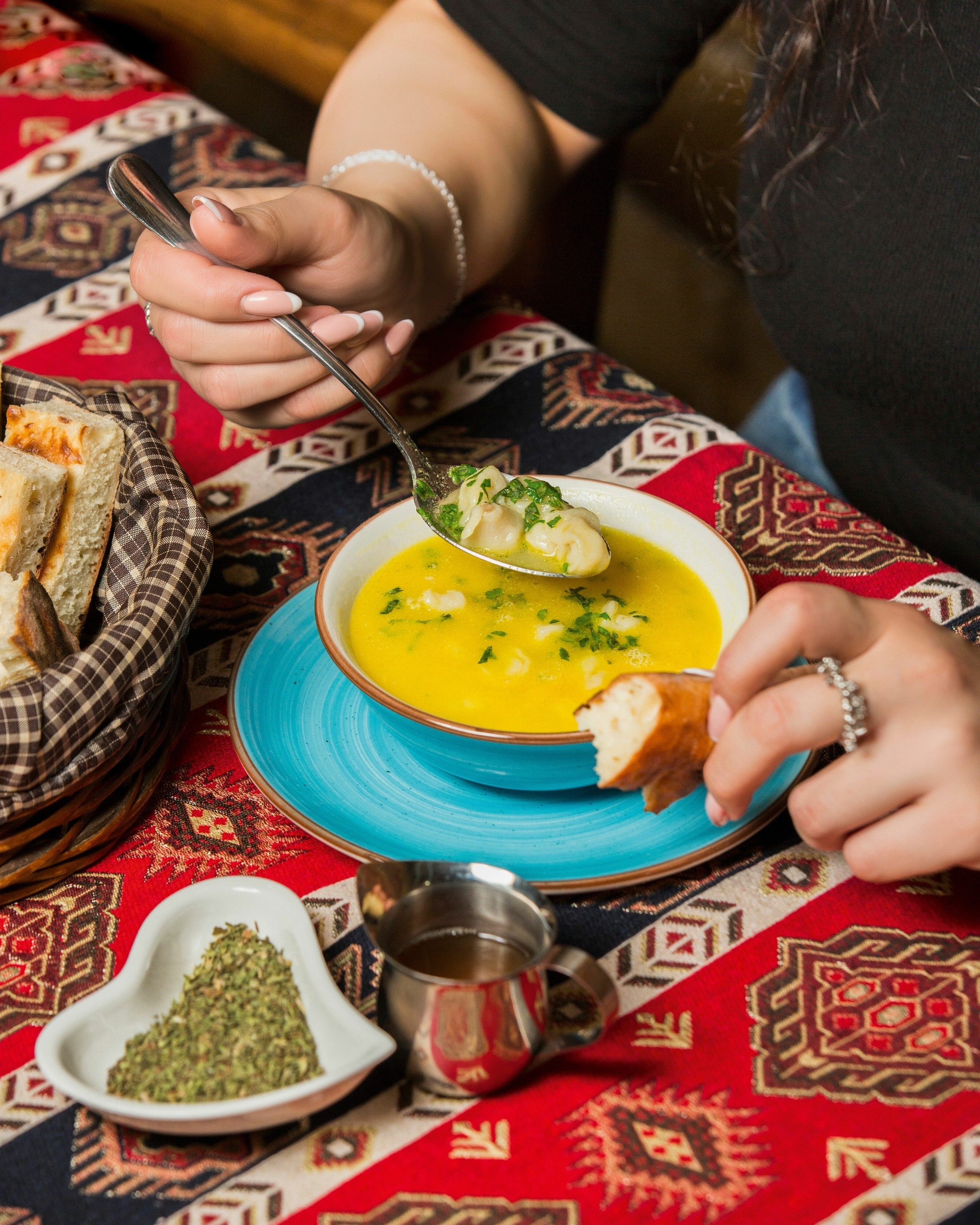 A person eating dushbara in a yellow sauce. A heart-shaped dish filled with herbs is on the table next to them.