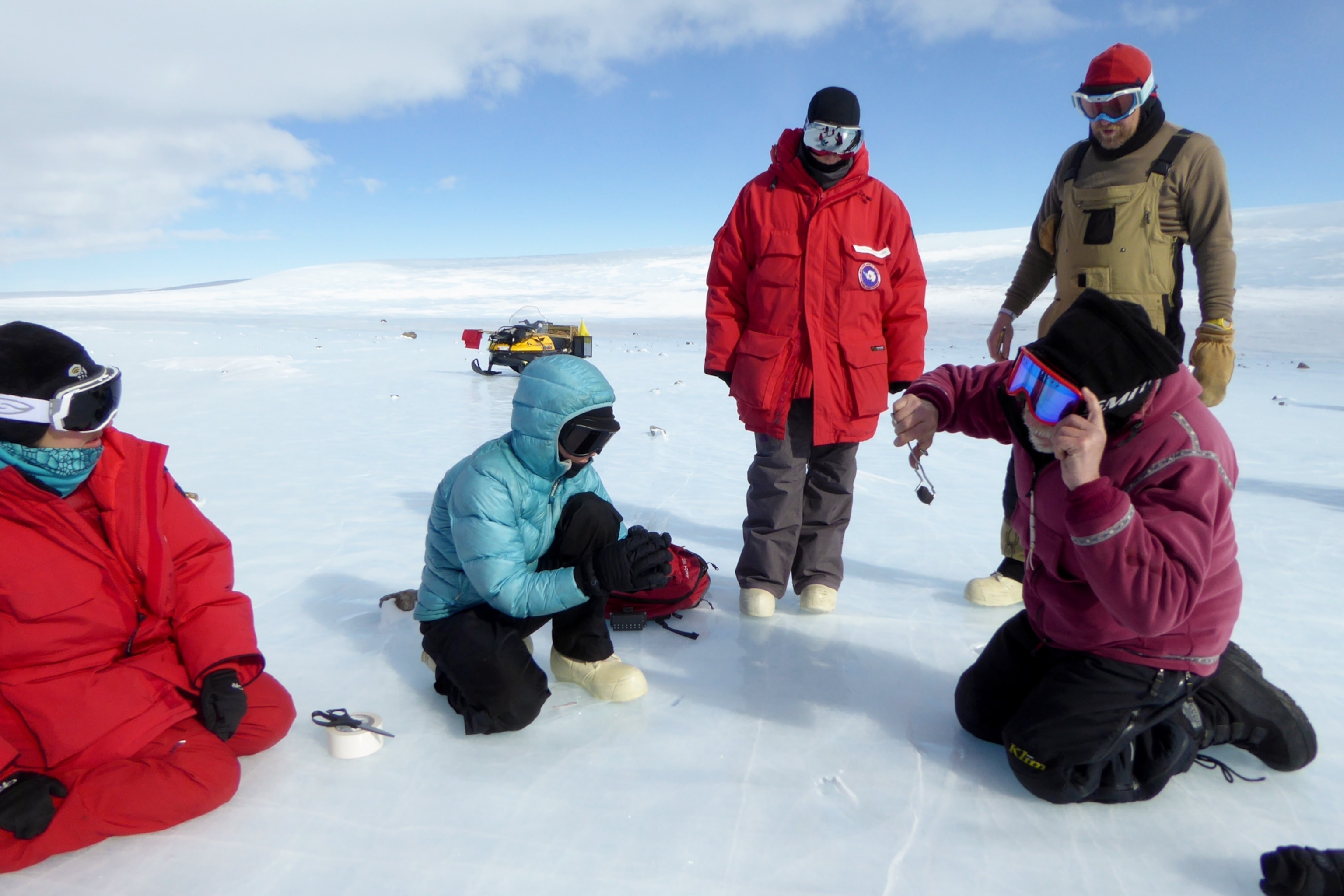 Members of NASA's ANSMET Antarctic expedition standing around a meteorite that one team member is holding with tongs.