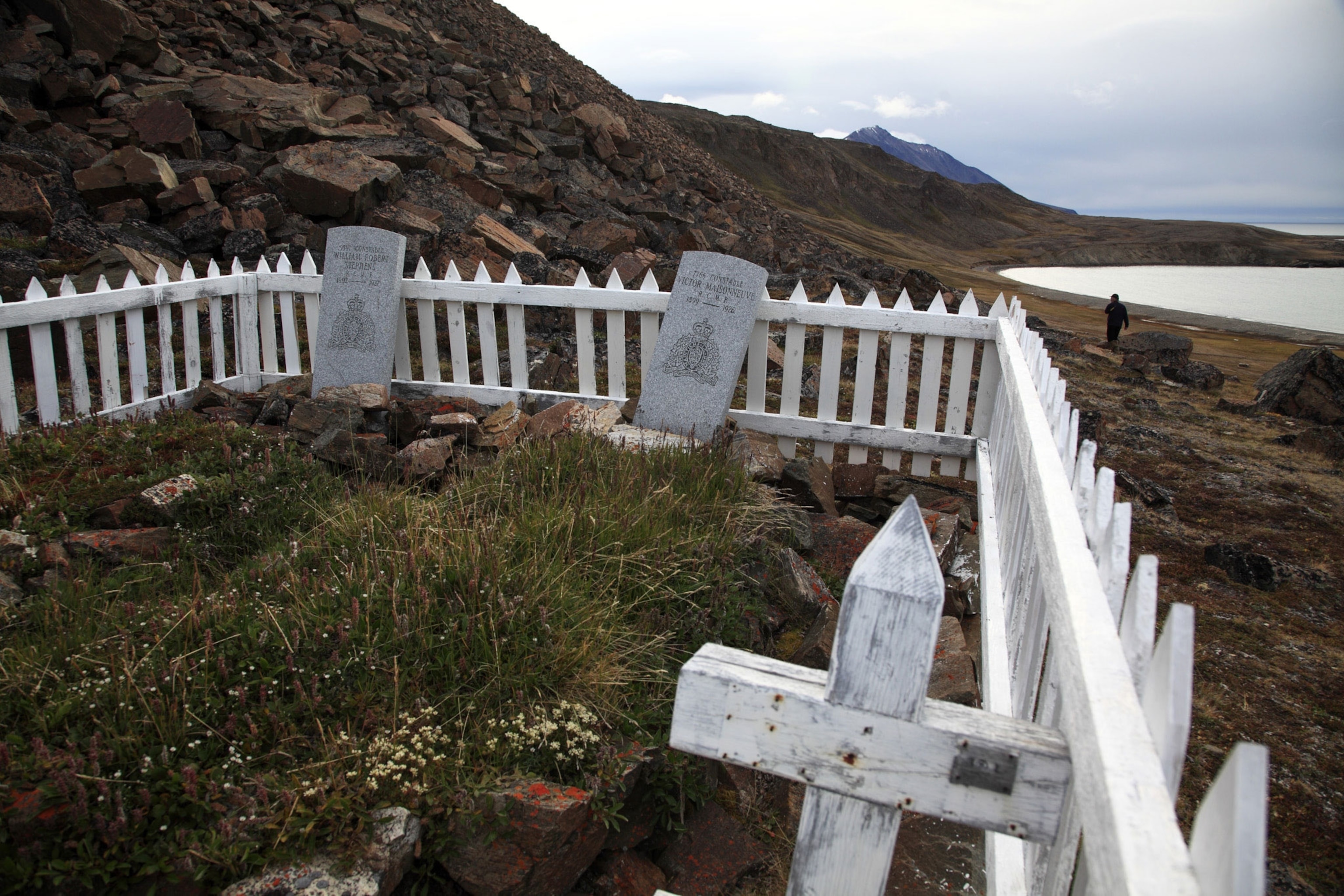 A small cemetery with only two tombs enclosed by a white fence sits at the bottom of a rocky cliff with mountains and ocean behind in the distance.