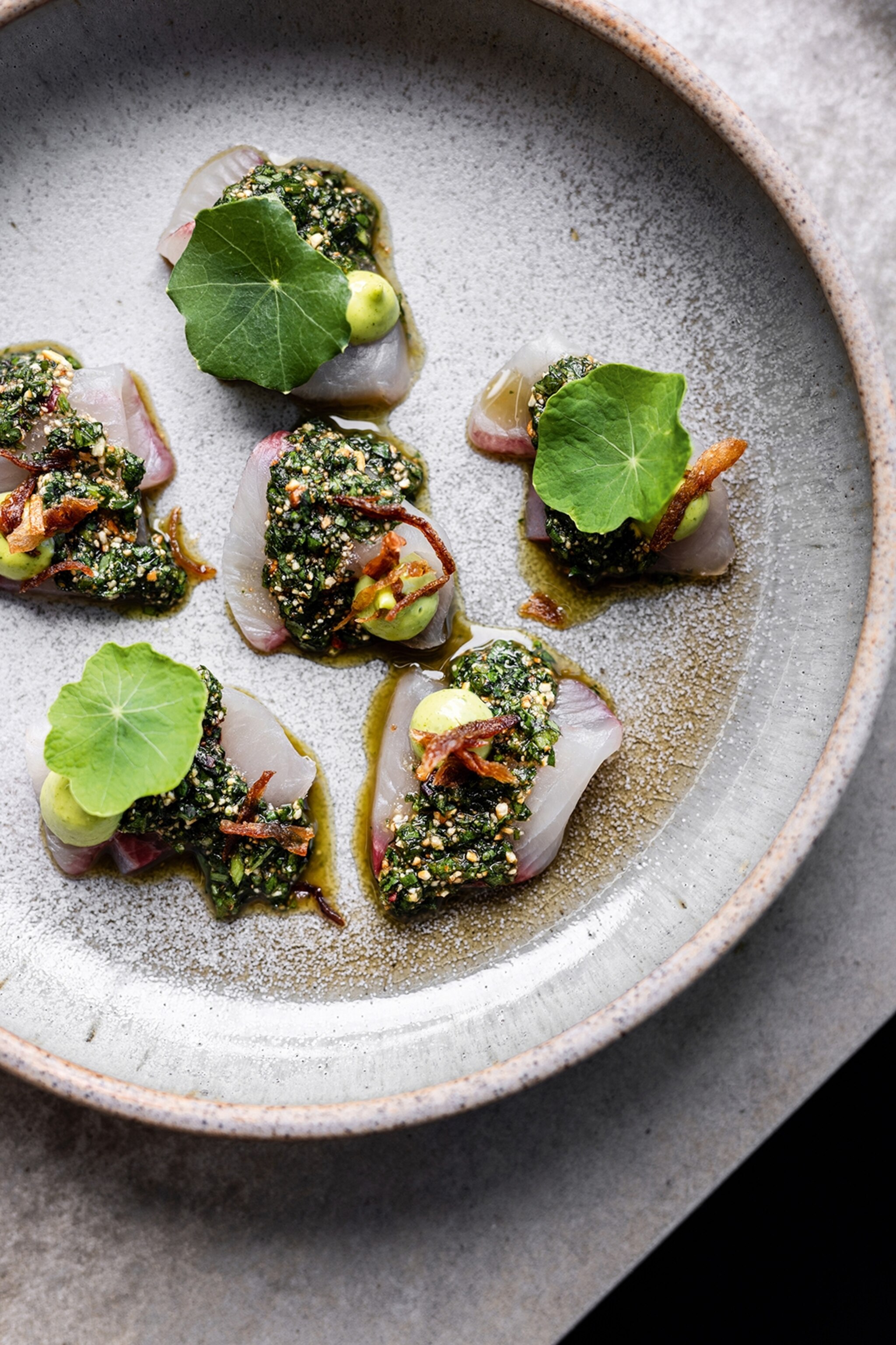 A plate of kingfish medaillons topped with an herby sauce, crispy flakes and leaves.
