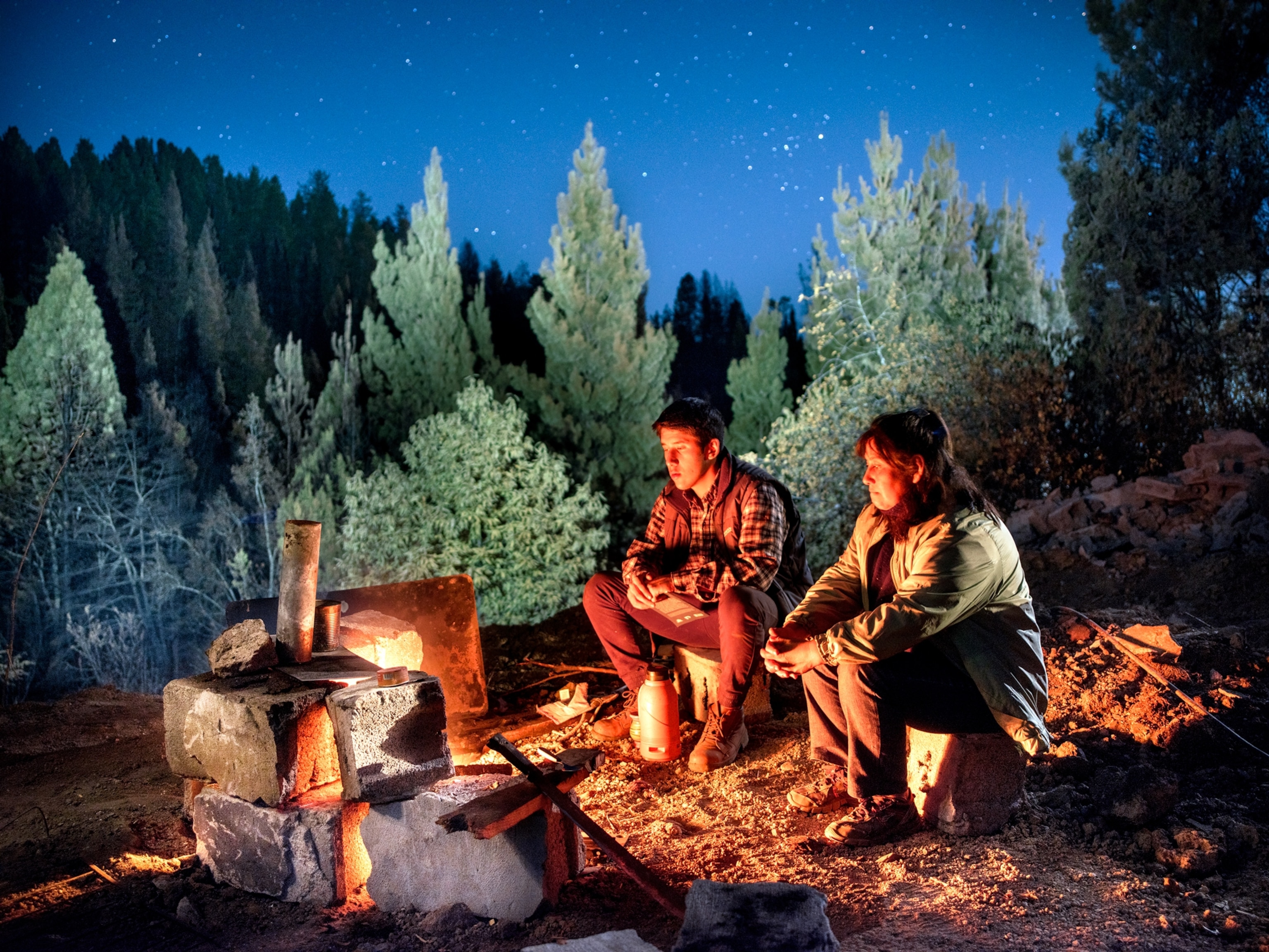 a man and woman sit near a small contained campfire
