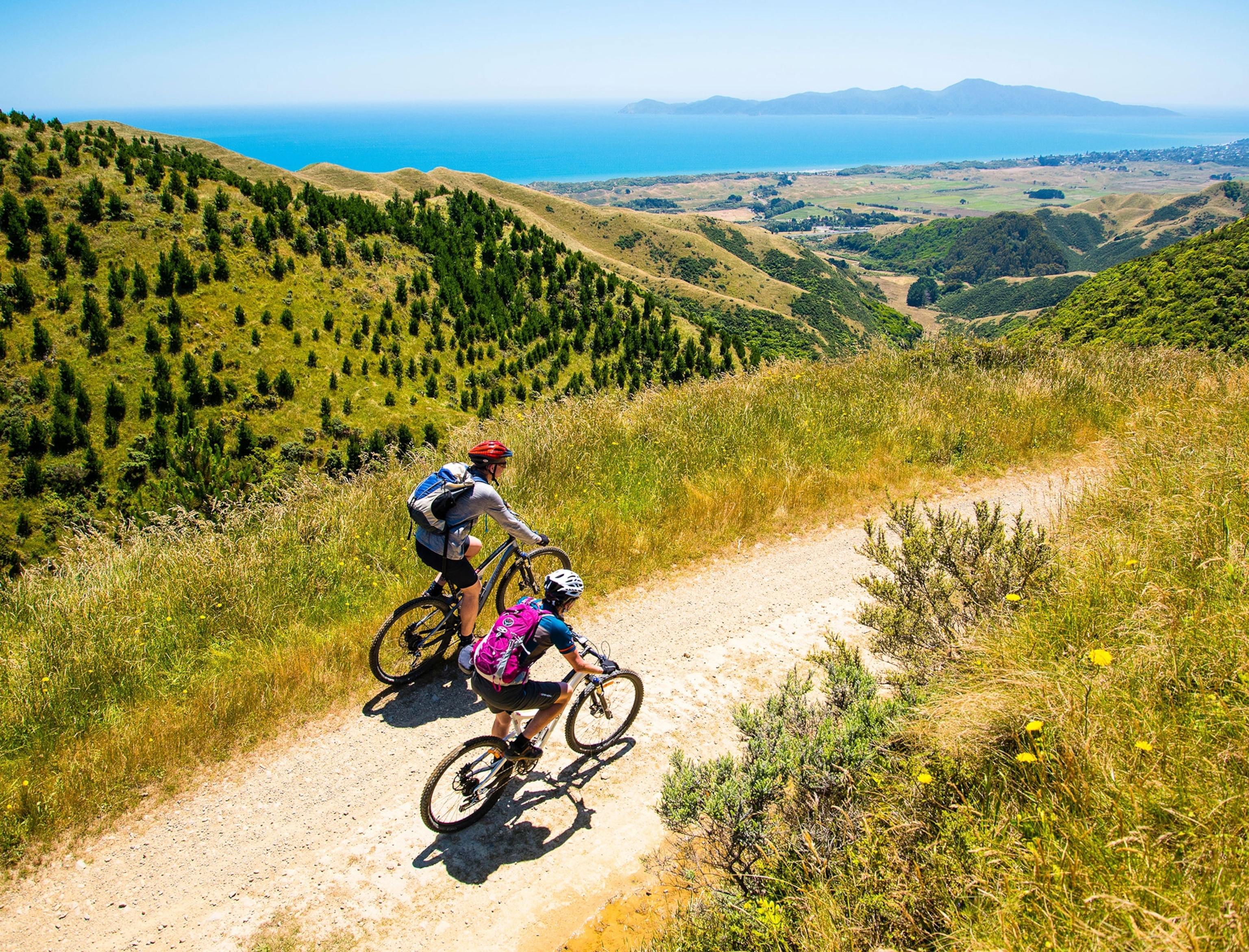 Two people riding bikes on a dirt road surrounded by green hills and blue water in the distance.