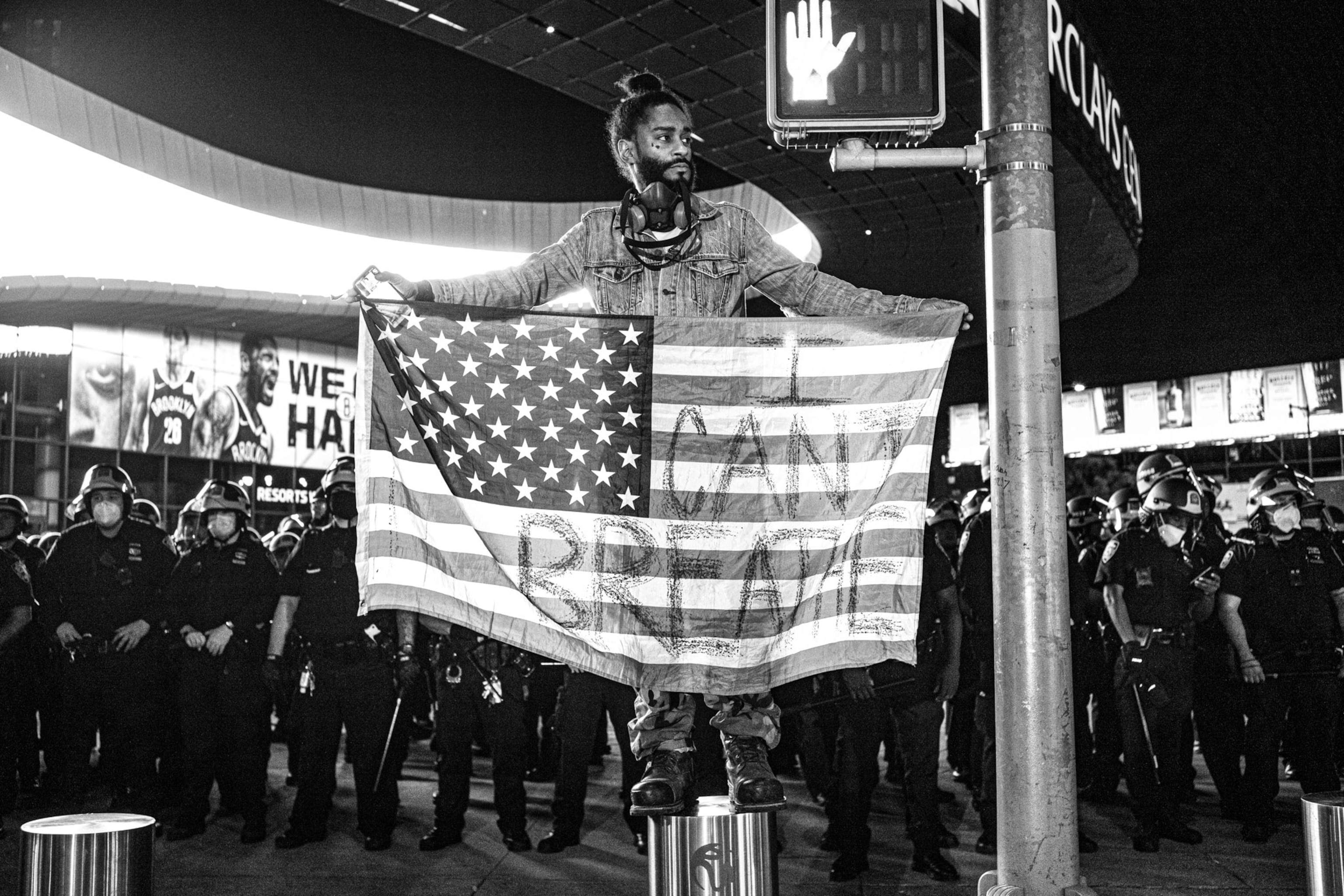 a person standing on street divider holding an American flag that reads "I can't breathe"