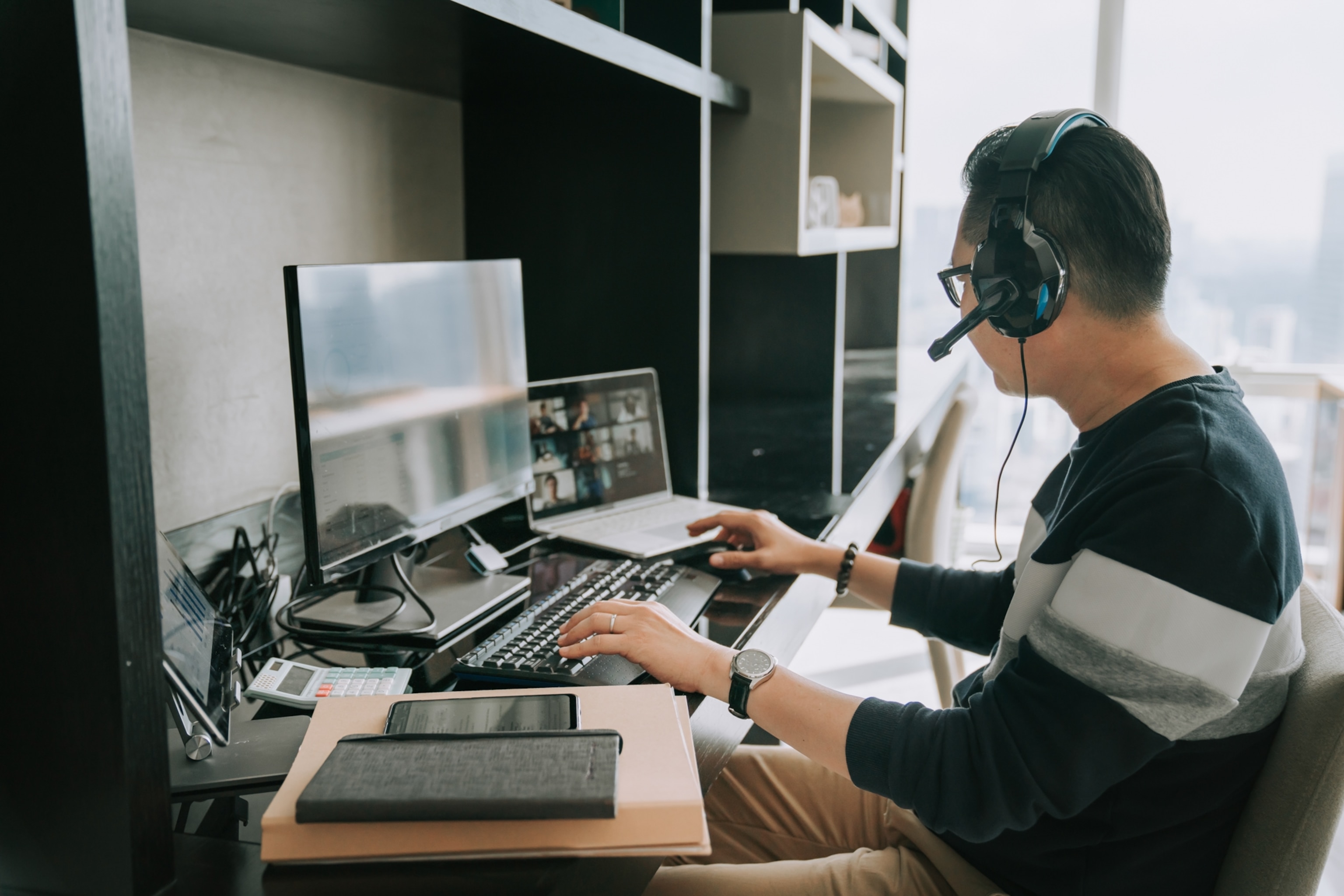 A man multitasking at his desk, using two monitors, a phone, and a headset.