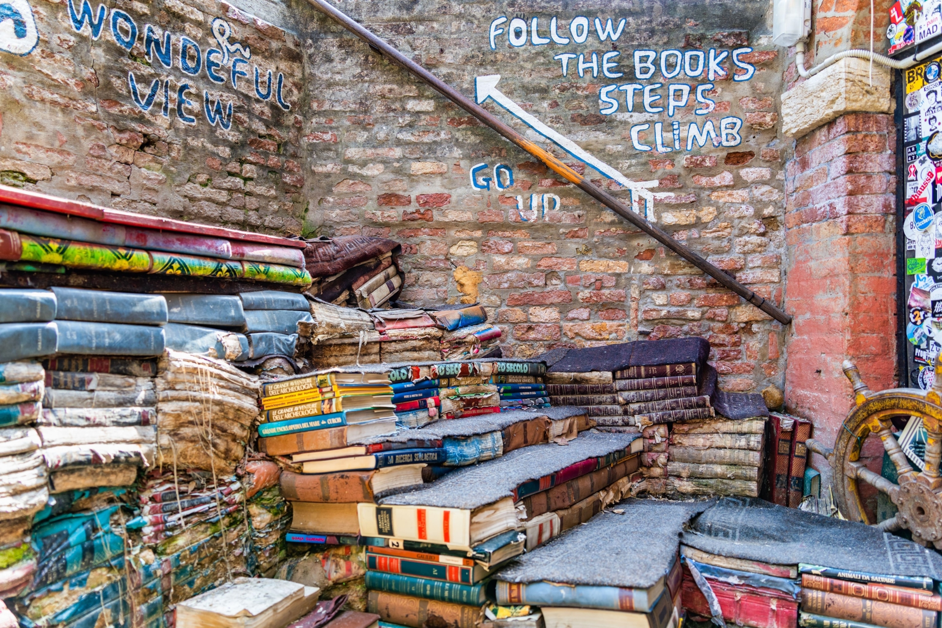 Stacks of books made into a short staircase against an old brick wall, where graffiti instructs visitors to, "Follow the booksteps," "Climb," "Go up." At the top of the book staircase more graffiti displays, "Wonderful view."