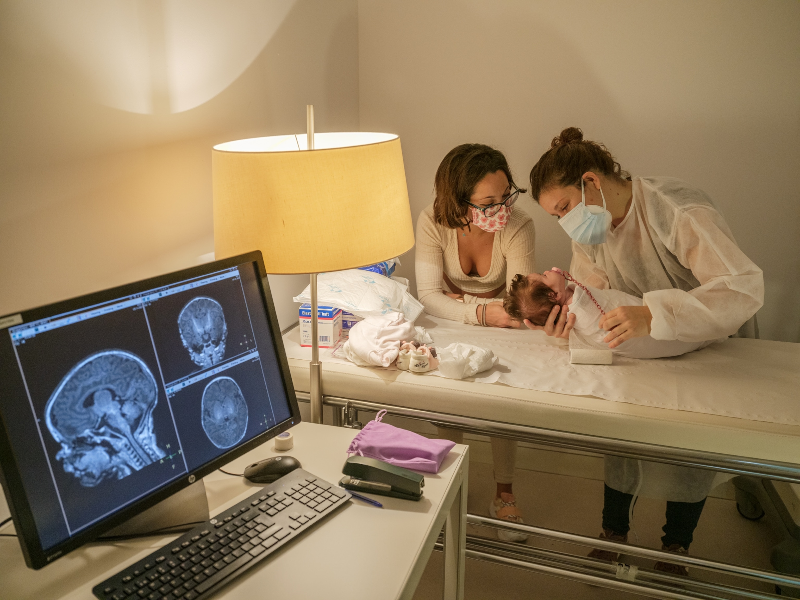 Two women tend to a young baby, a brain scan is on a computer screen in the foreground