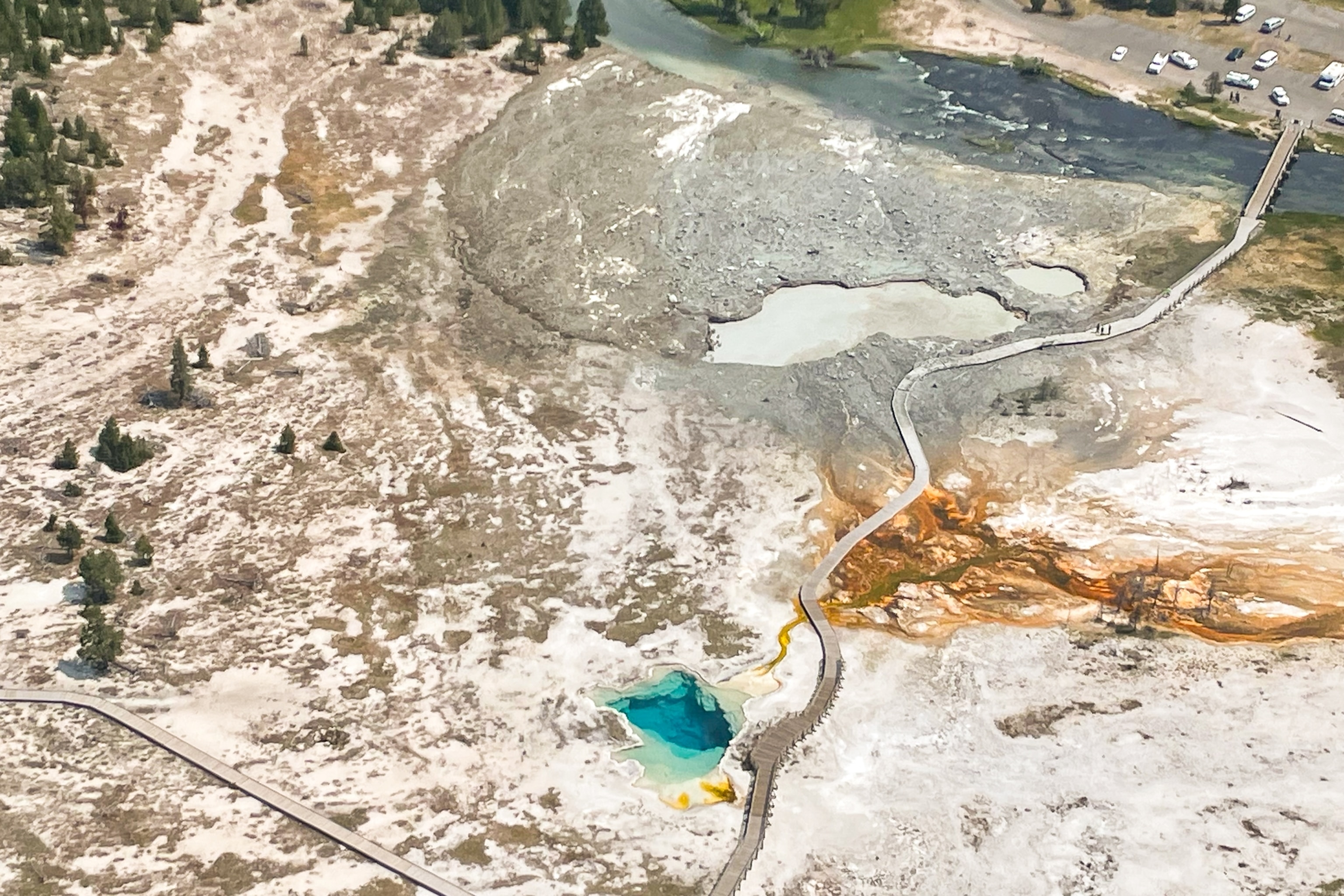 An aerial view of Biscuit Basin. One geyser basin towards the bottom of the photo is still turquoise blue. Further above, the Biscuit geyser basin is a murky grey, and the adjacent boardwalk is covered in rubble.
