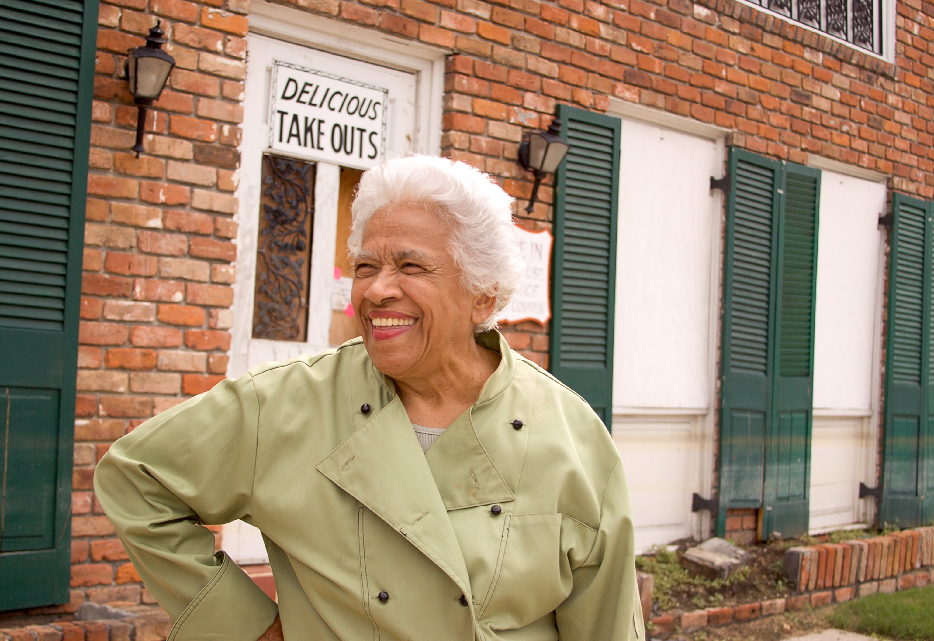 An elderly woman in a light lime green coat stands outside a brick building with white windows and green shutters. A sign above her says "Delicious Take Outs."