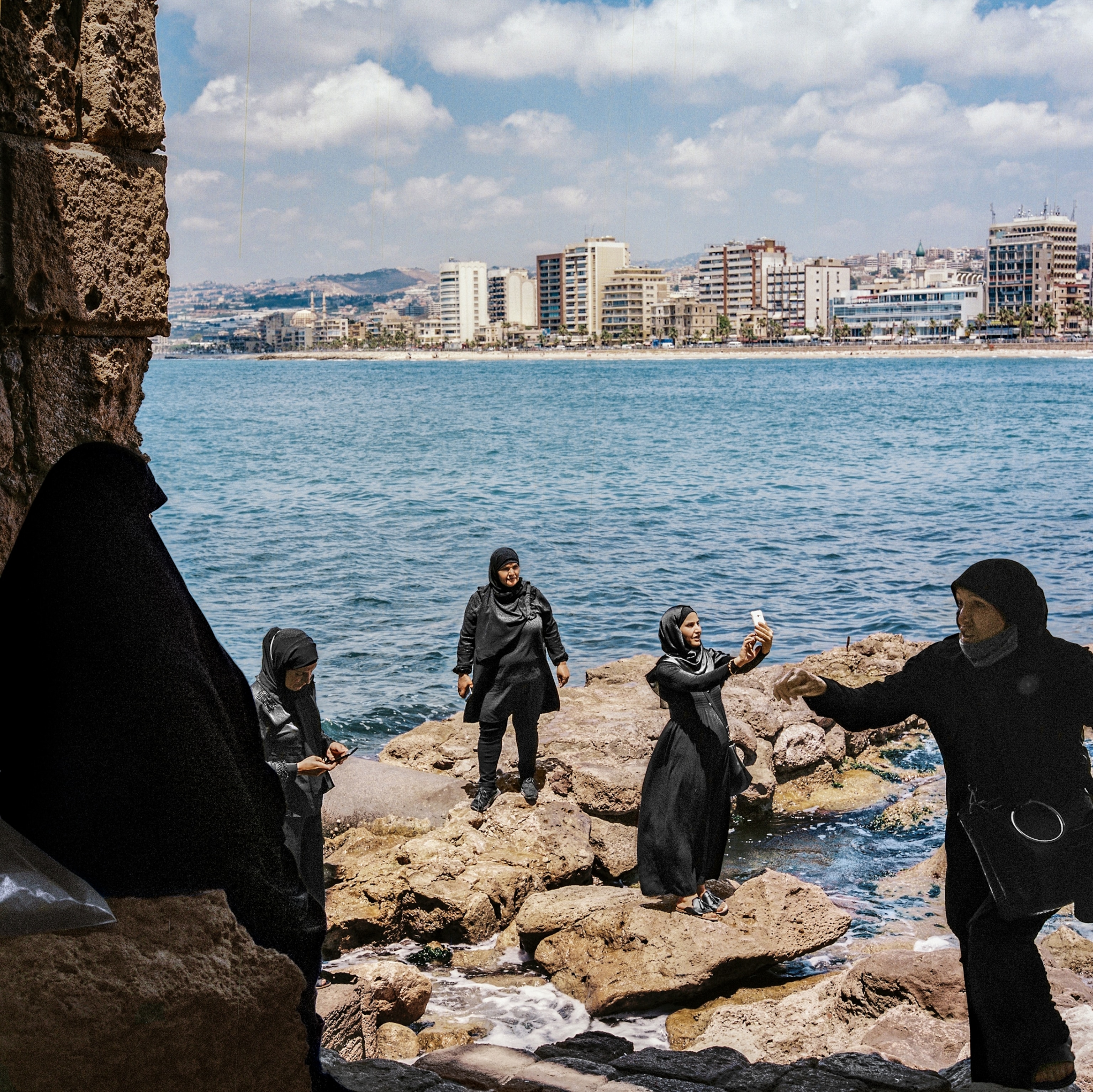 Picture of 5 women in black clothing stand dispersed on rocky shoreline with a cityscape behind them. One woman is taking a selfie with her phone.