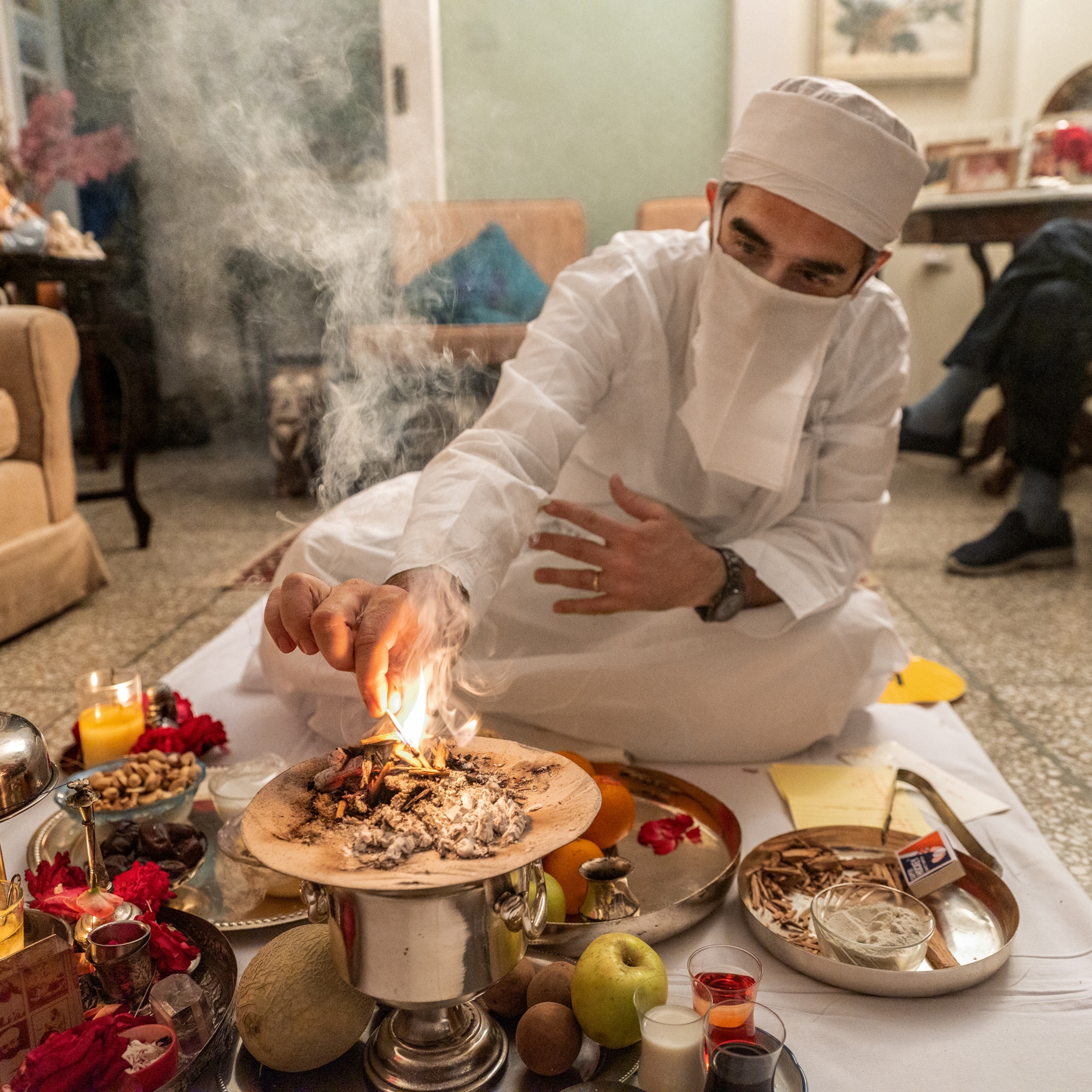 A priest wearing all white, is seated criss cross in front of ceremonial objects holding a a lit match.