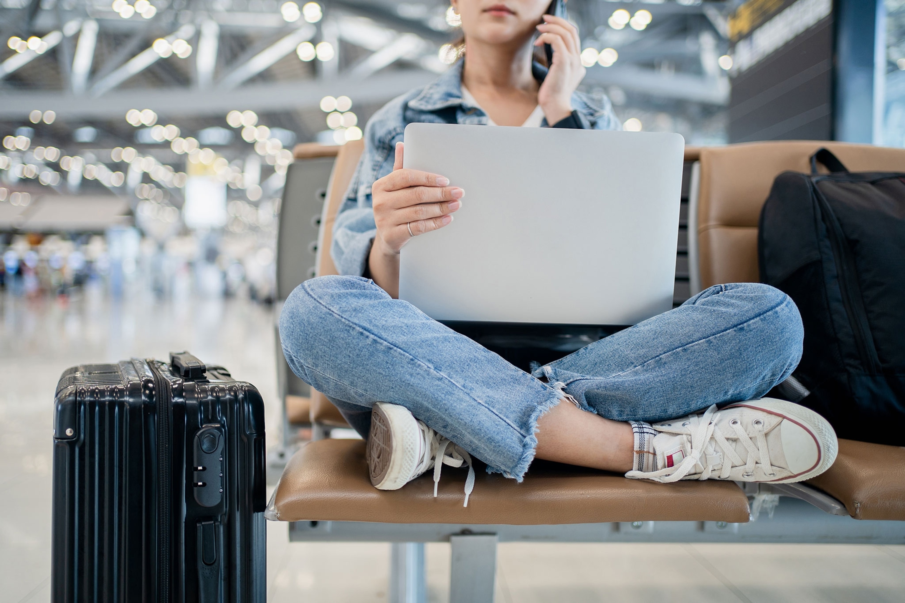 Woman sitting with her legs crossed while working on a laptop and talking on a cellphone with a suitcase to the left of her in the frame.