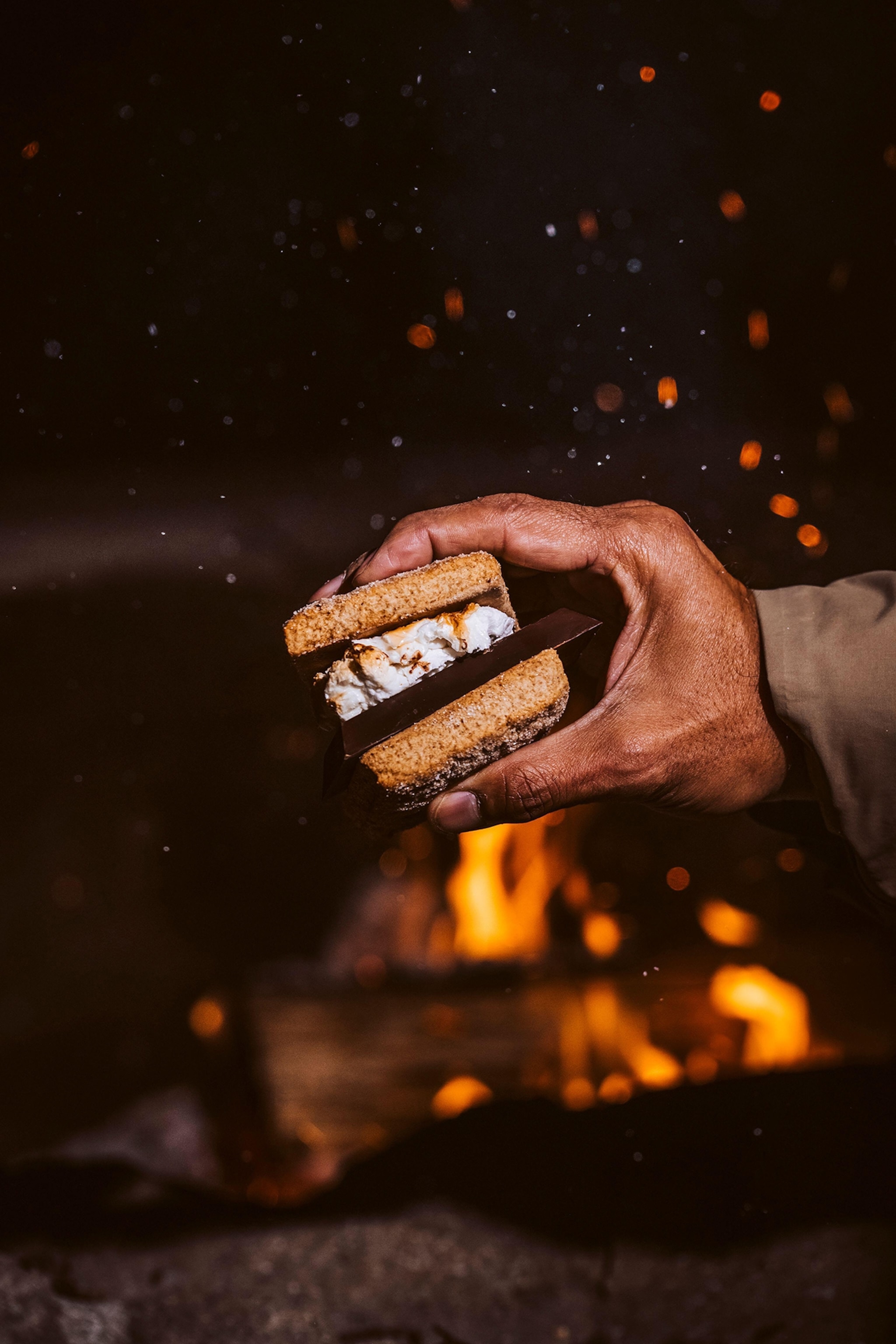 Man's hand holding a s'more sandwich with fire pit in background