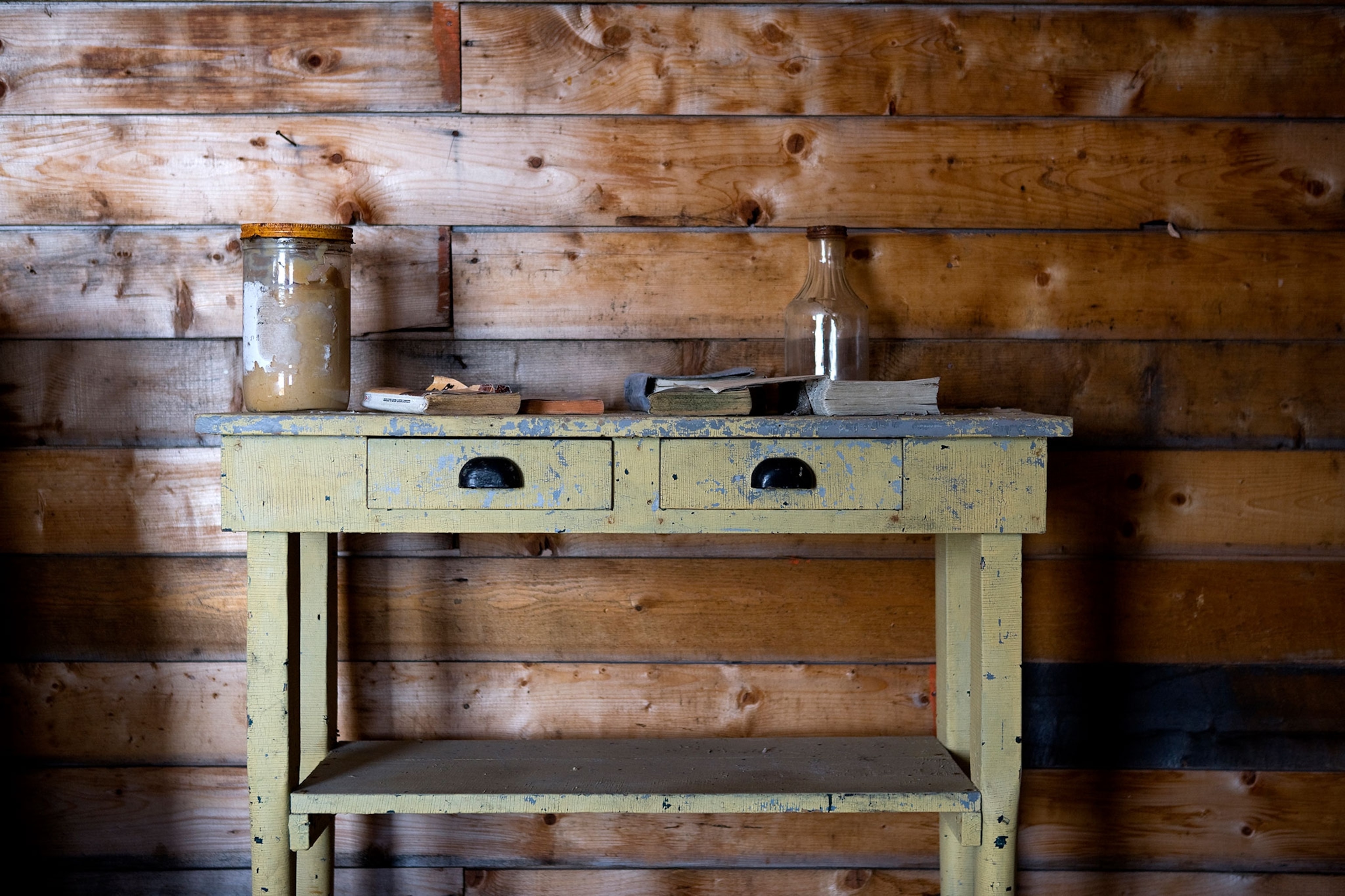 A green wooden table holding miscellaneous books and jars placed against a wooden wall.