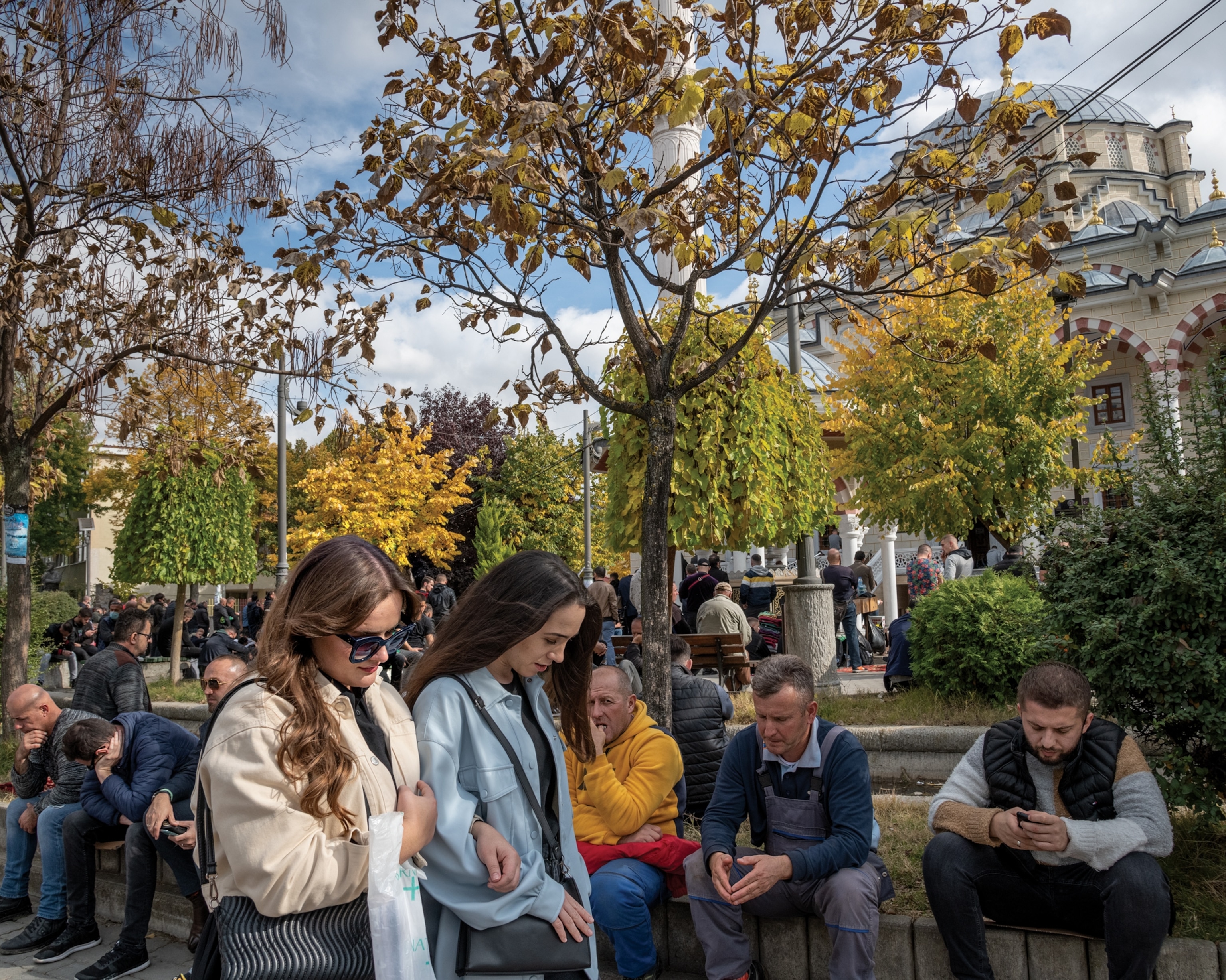 Picture of two women walking in front a many men sitting on a concrete ledge in a crowded park.