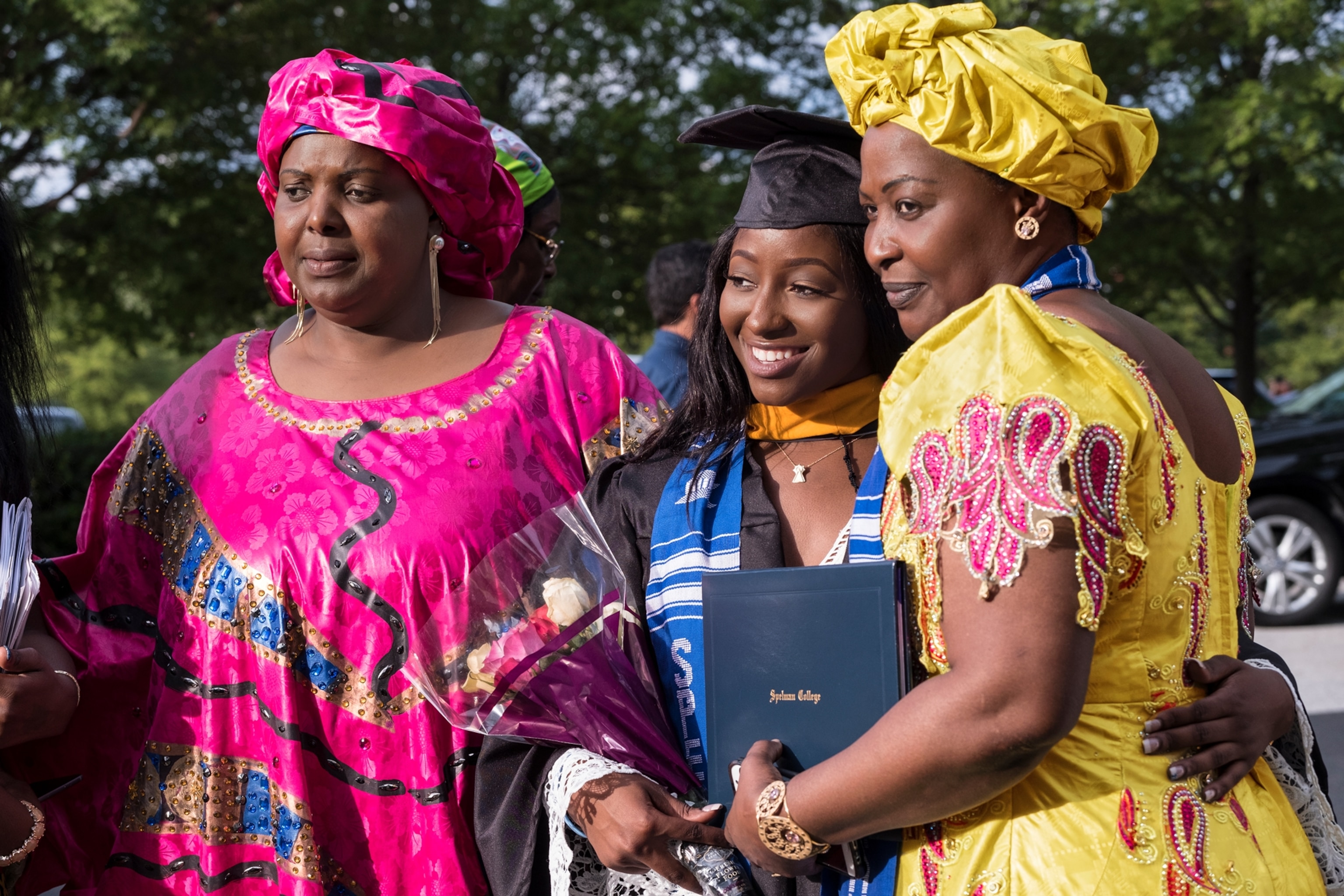 two African-American women in traditional dresses embracing another one in graduation gown.