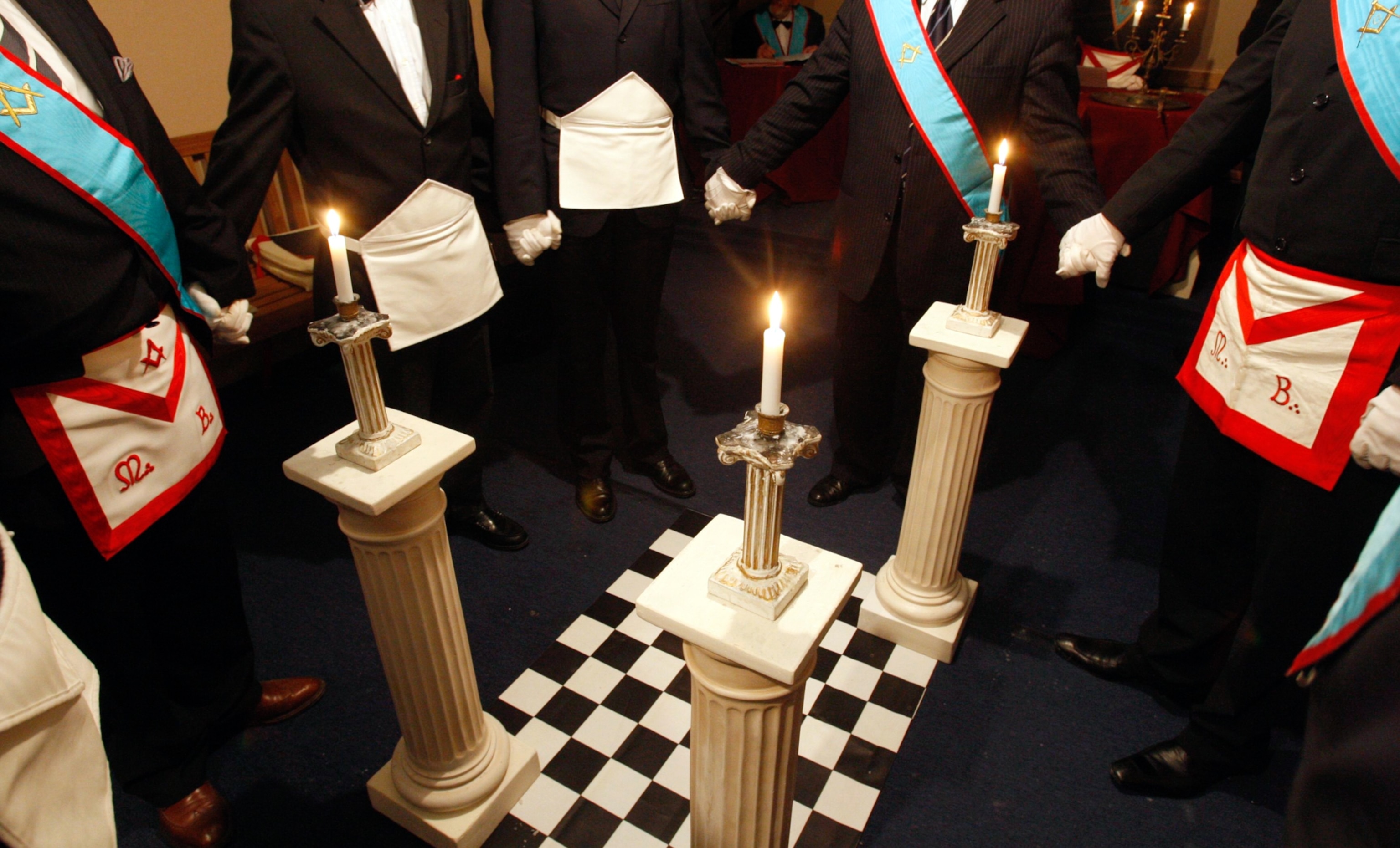 Freemasons standing around three candles on columns placed at three corners of a rectangular checker-board pattern platform.
