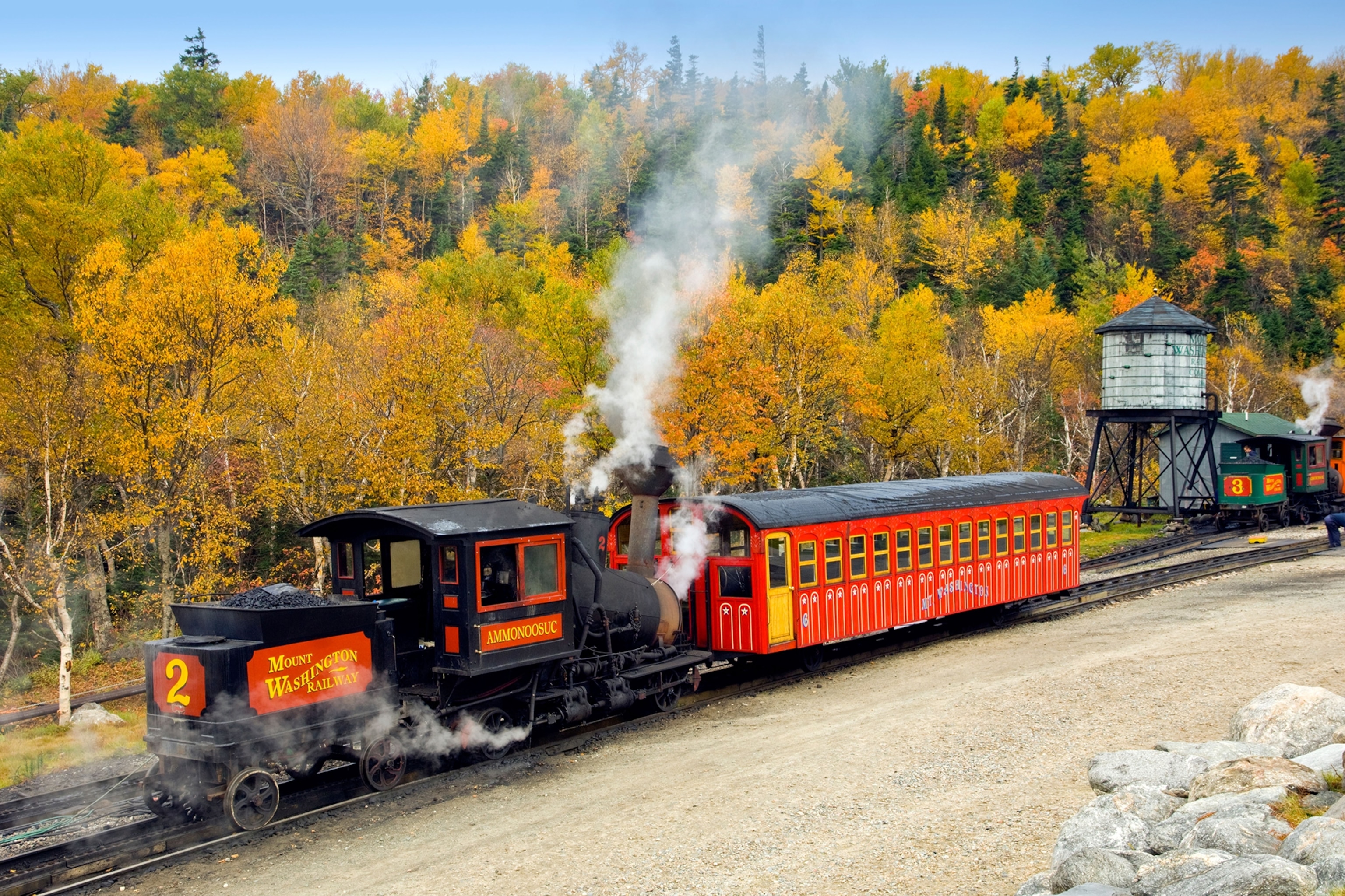 Red historic steam railway running along tracks surrounded by orange and yellow autumnal trees