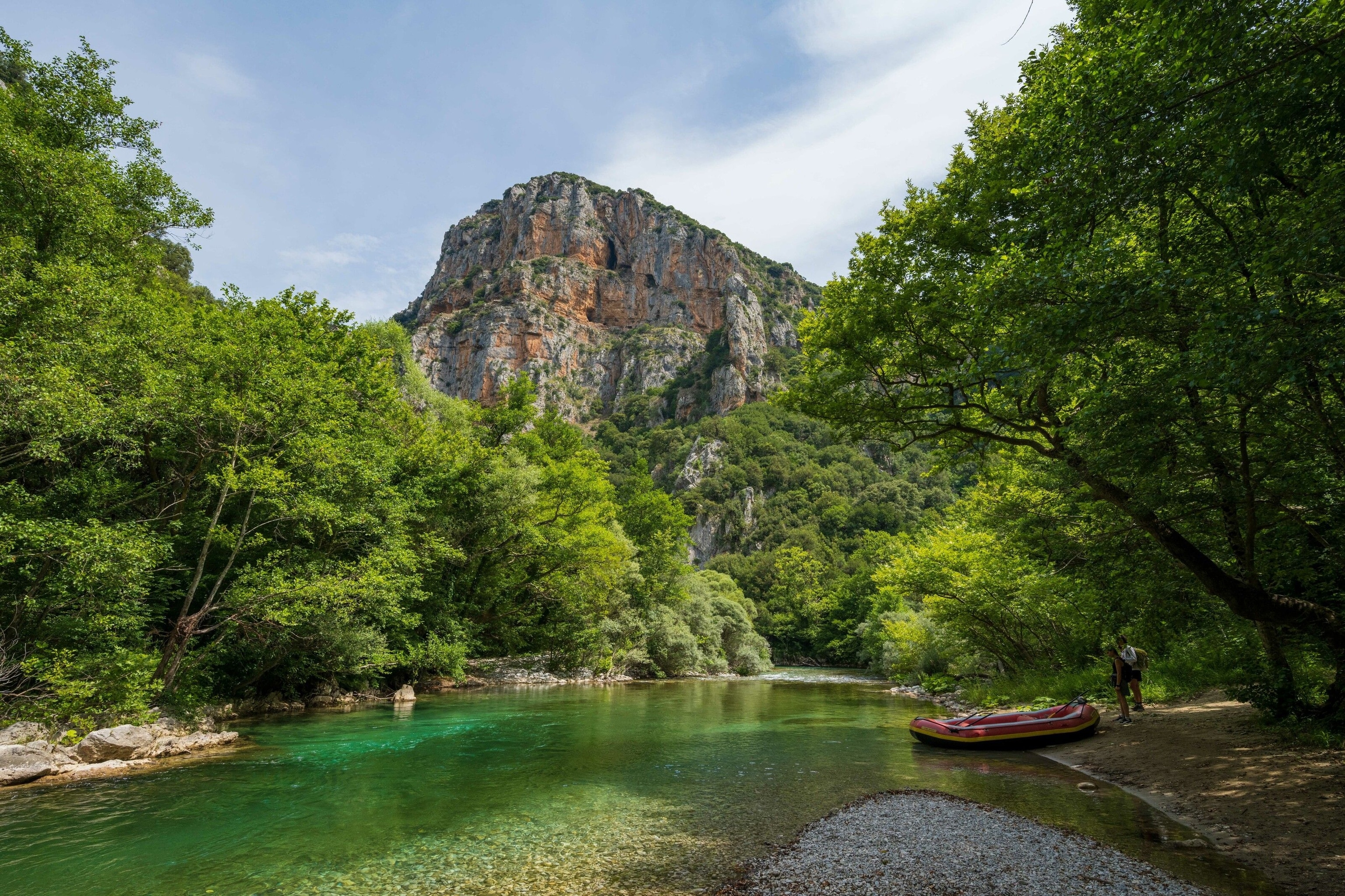 Voidomatis river. It is still and looks shallow. A red tender is on the right bank. Two people look at it. The water is green.
