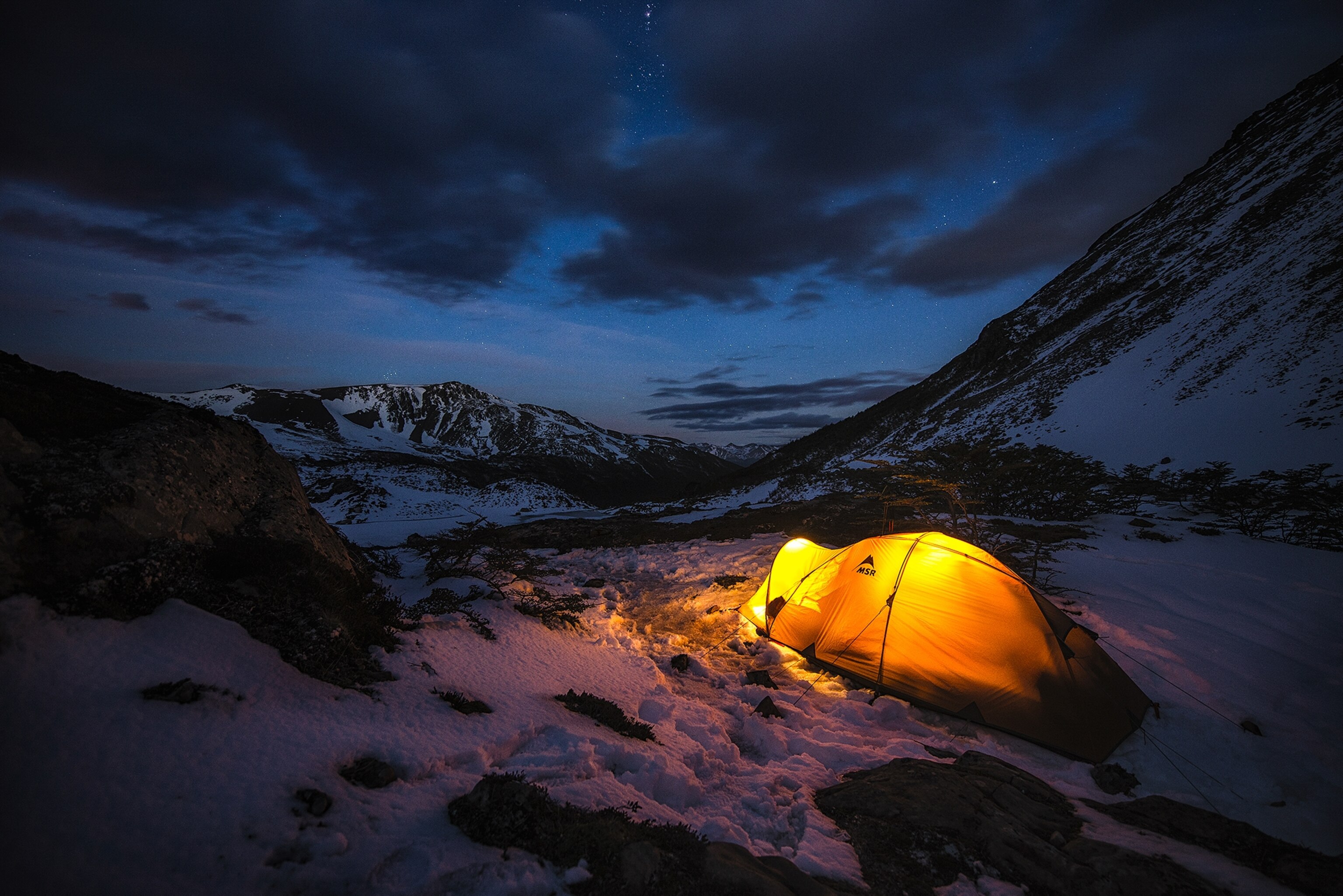 A tent casts a golden glow across snowy and rocky terrain, as stars peak through breaks in the night clouds above.