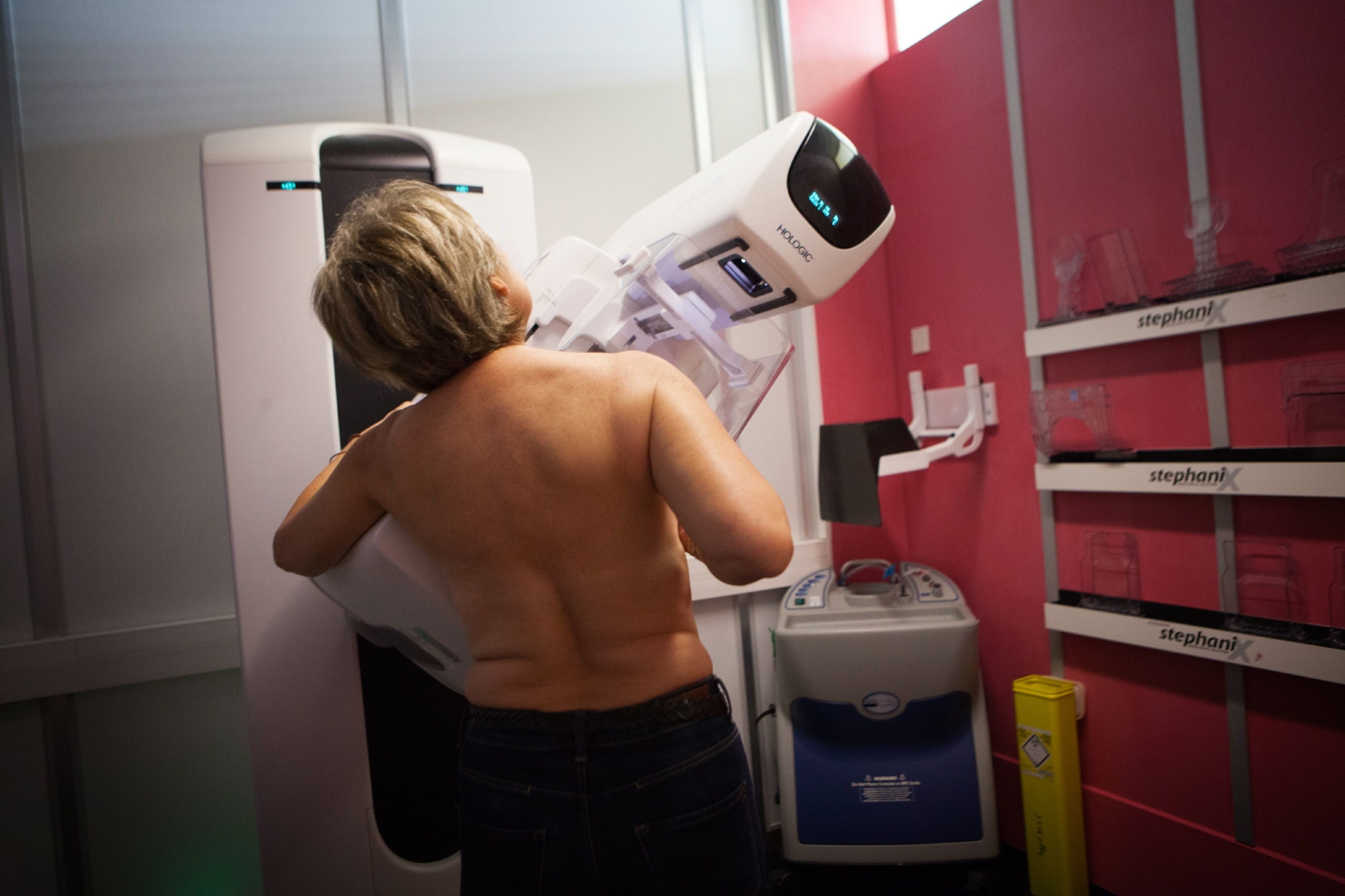 A woman seen from behind with her shirt off as she leans with a mammogram machine.