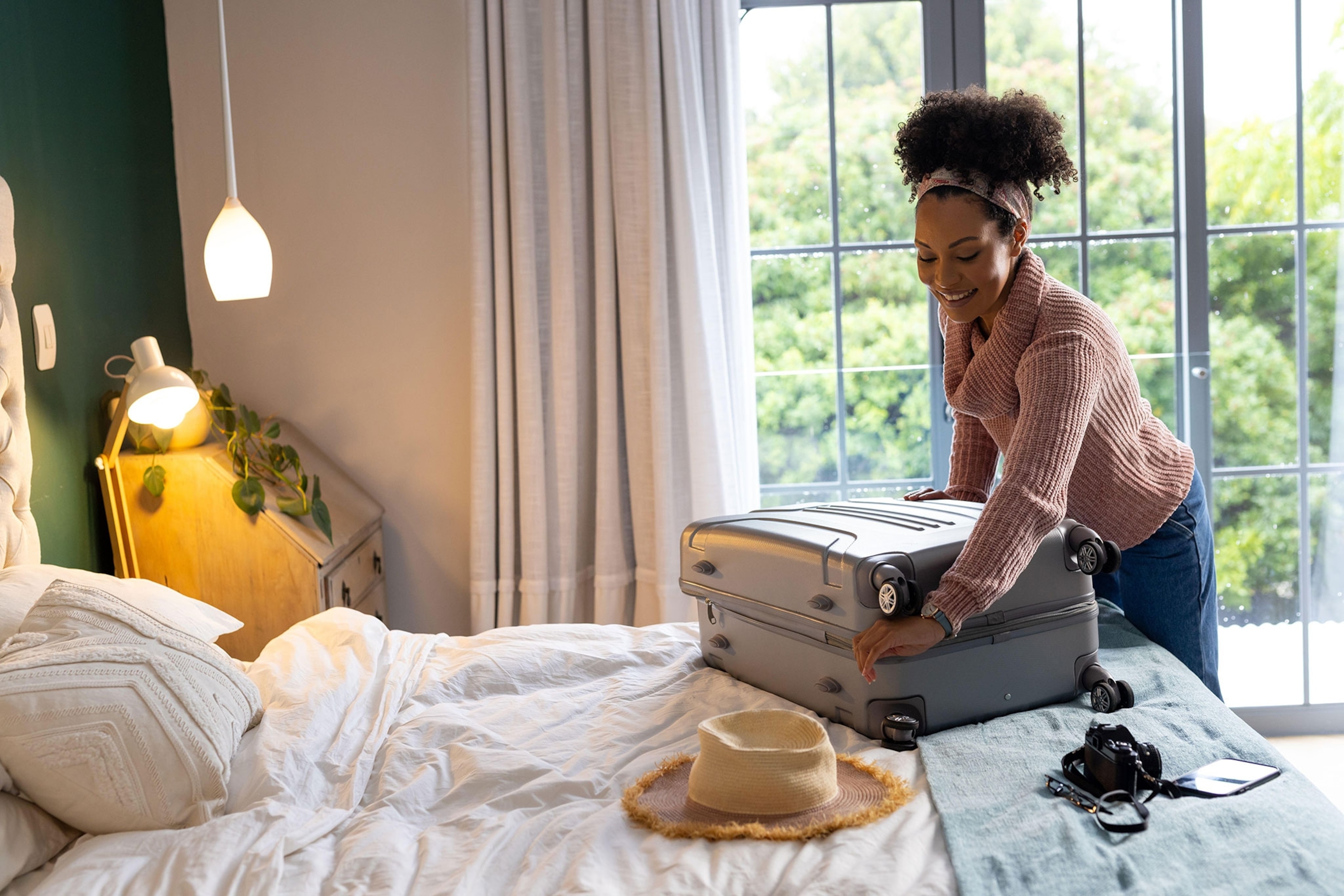 A woman packing a suitcase on a bed with lamps in corner of the room and windows in the background.