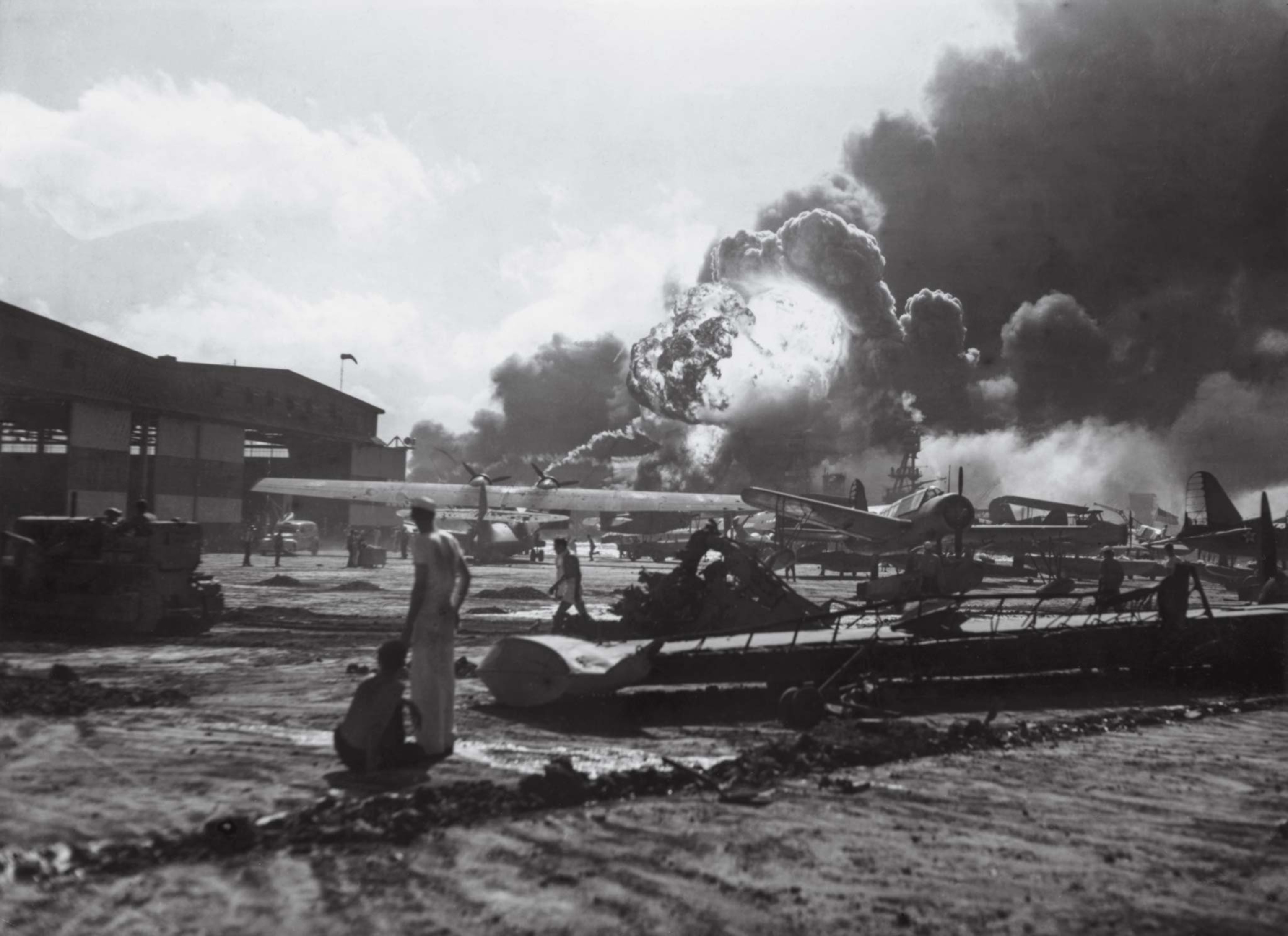 A sailor observes explosions and stands amid wreckage at Ford Island Naval Air Station during the Japanese attack on Pearl Harbor.