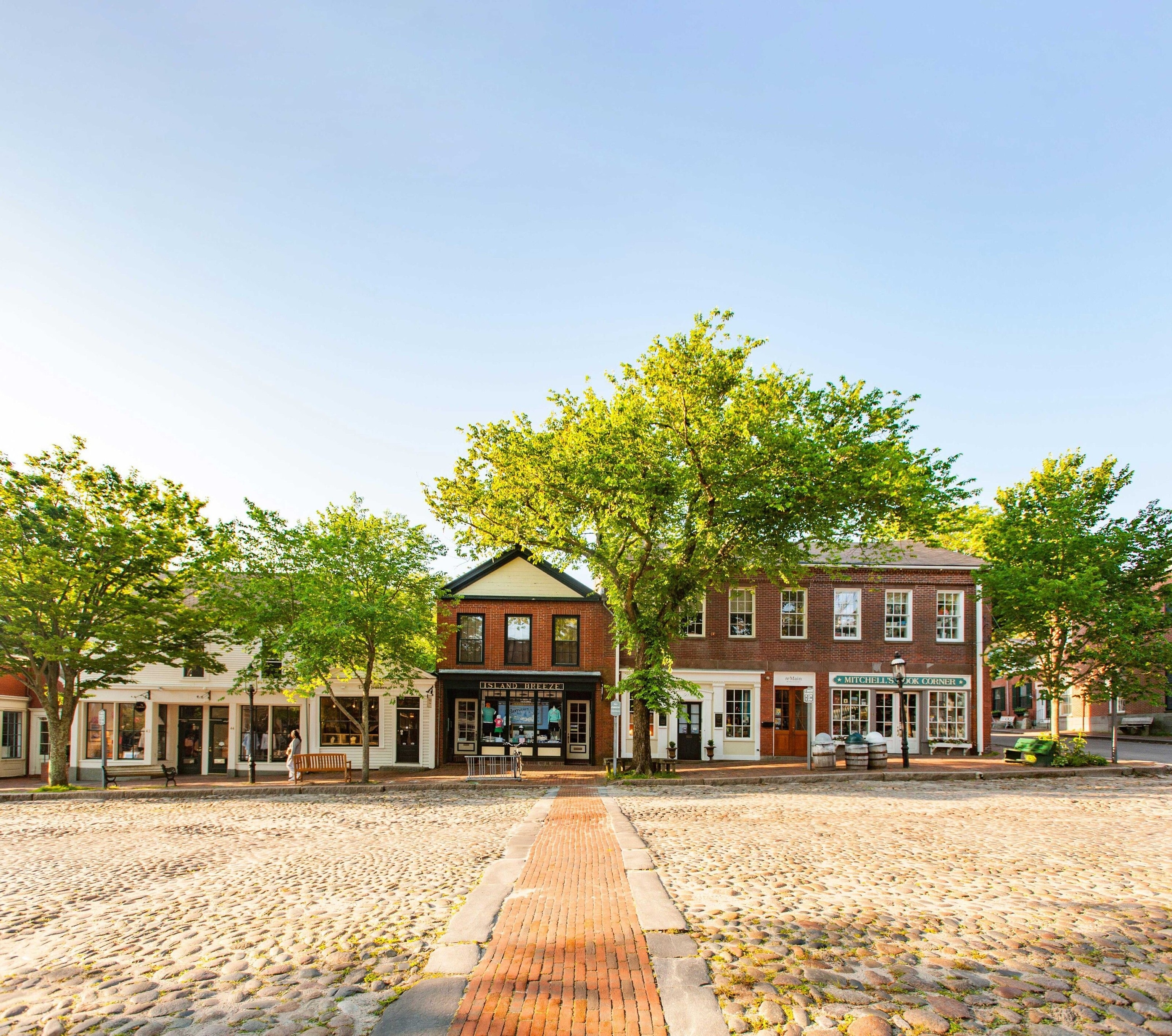 The cobbled street of Nantucket. There is a small path of orange brick, which leads to quaint stores tucked behind tall green trees.