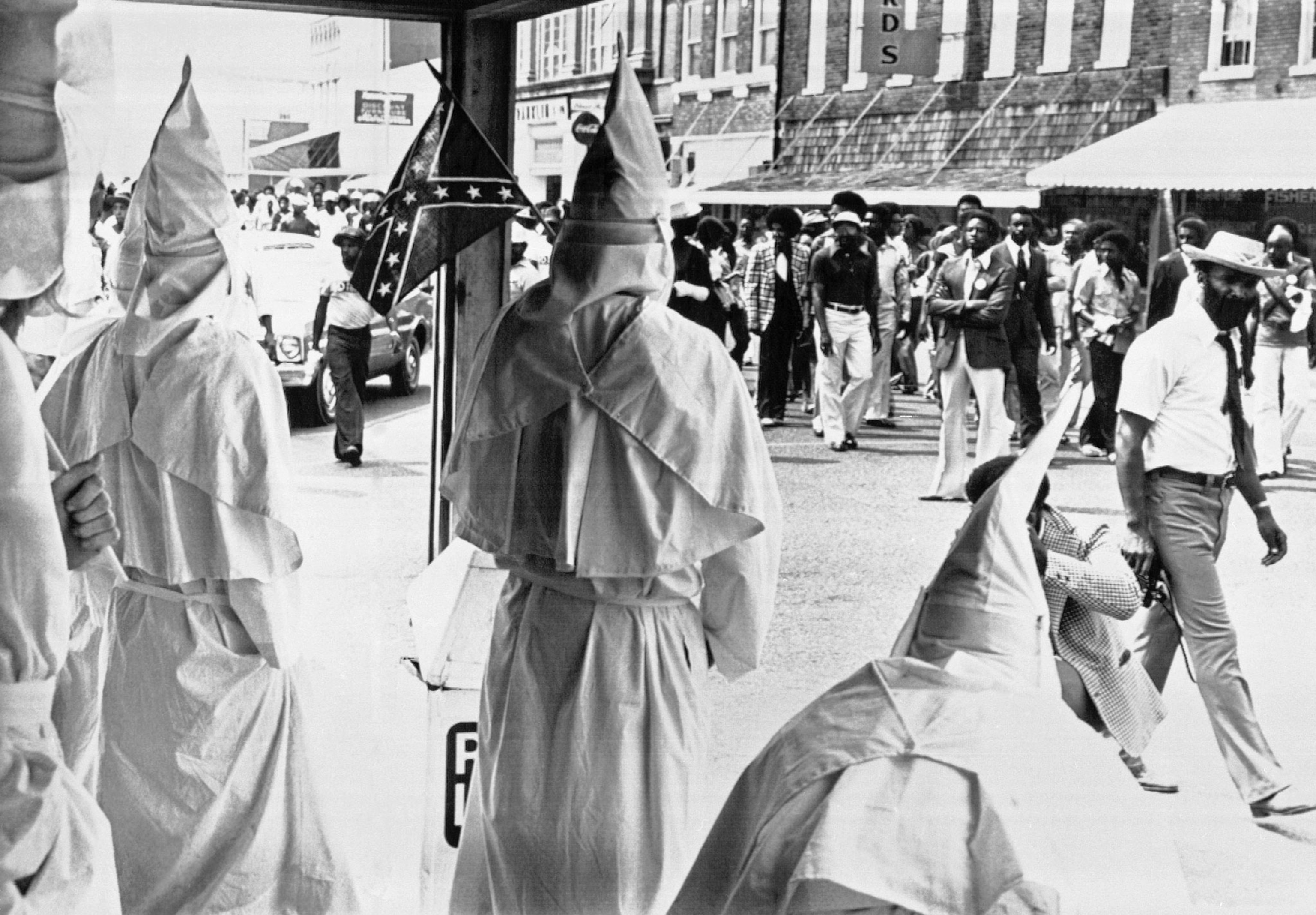 African Americans walk down a street while men in ku klux klansmen watch