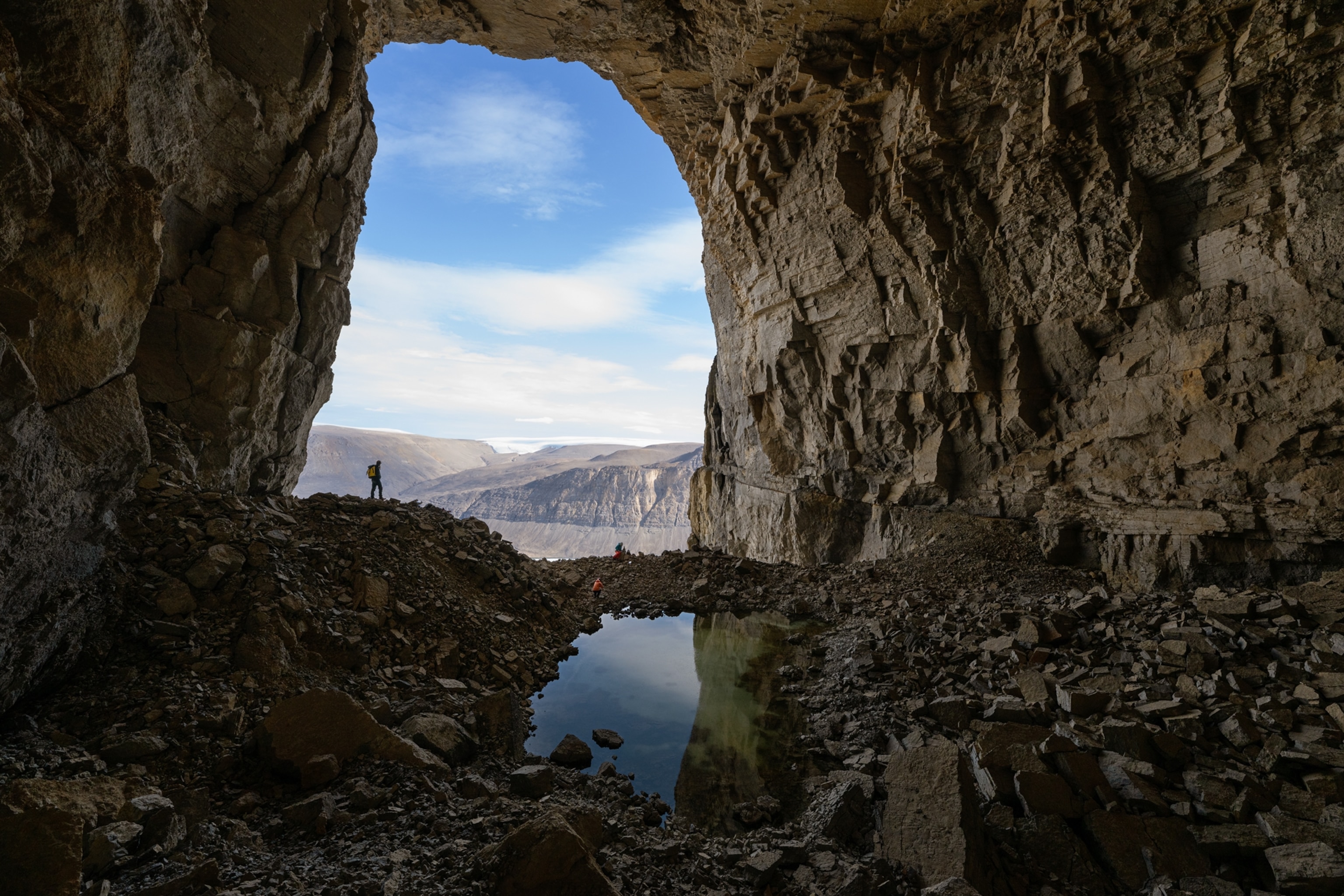 Gina Moseley stands at the arch-shaped entrance of the cave. A small lake inside the cave is in the forground of the frame.