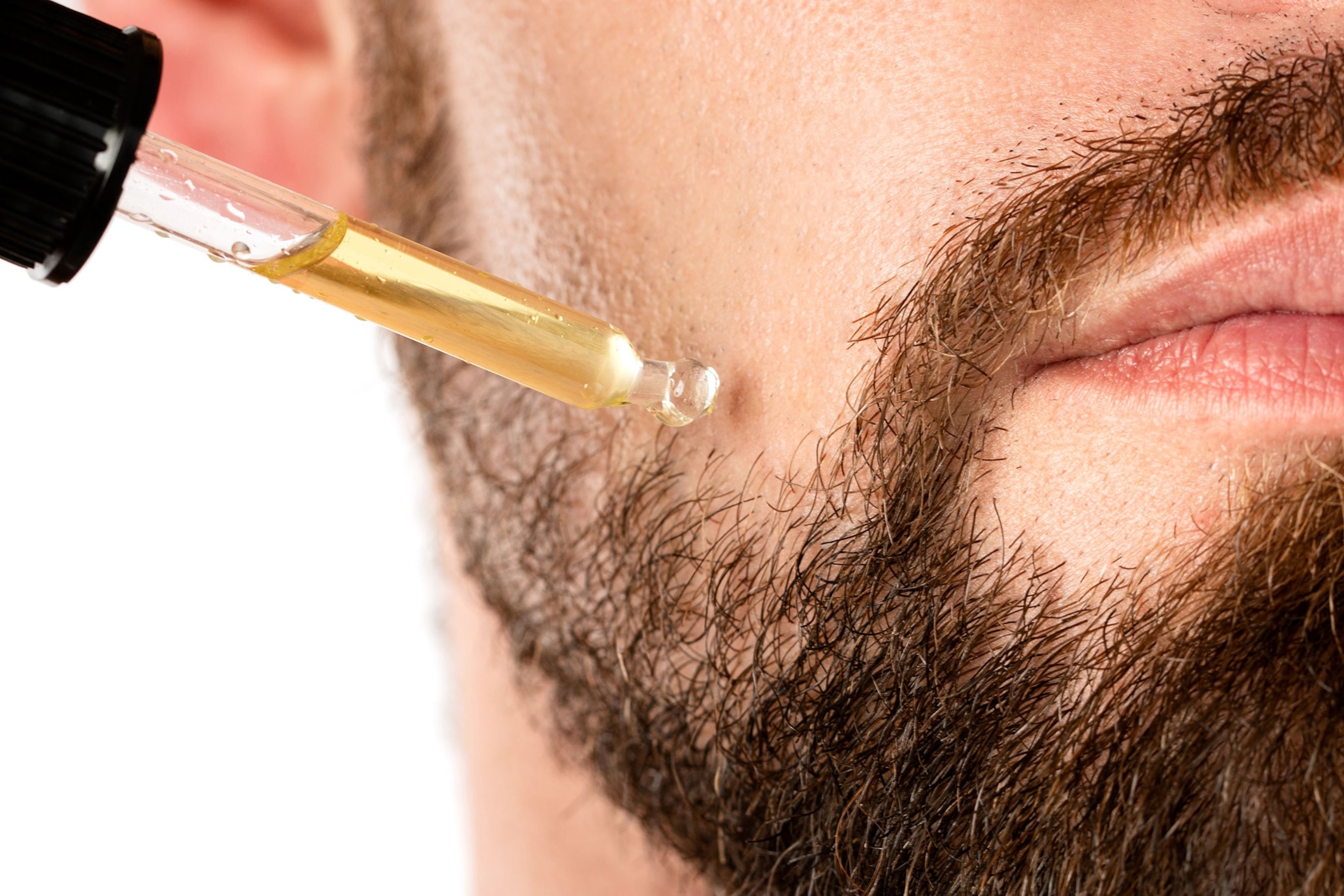 An eye dropper, with a gold liquid inside, being placed next to the brown beard line of a man