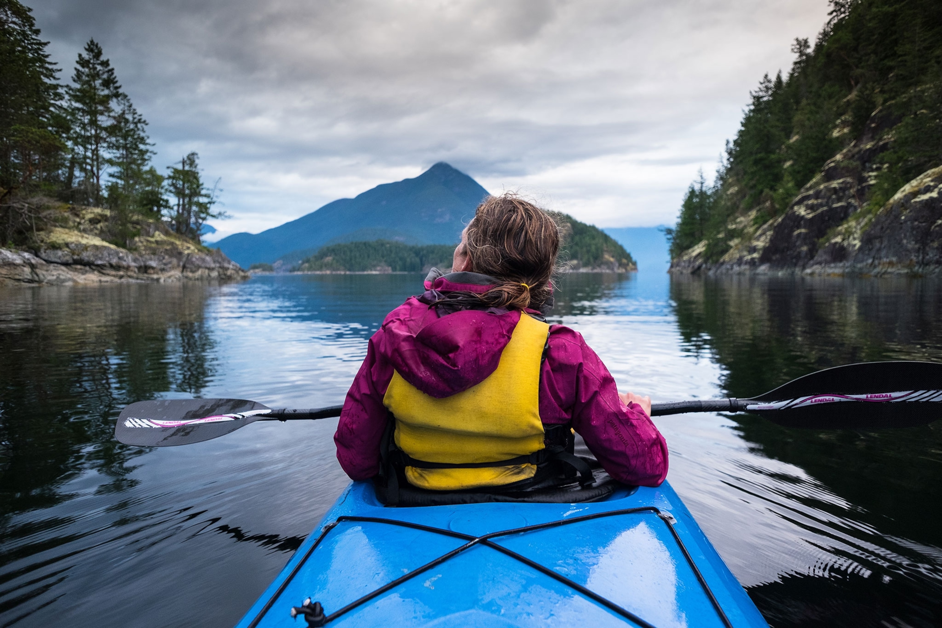 A woman with a mangenta rain coat, yellow life vest, paddling in a blue canoe with cliffs and trees to her left and right and a mountain straight ahead.