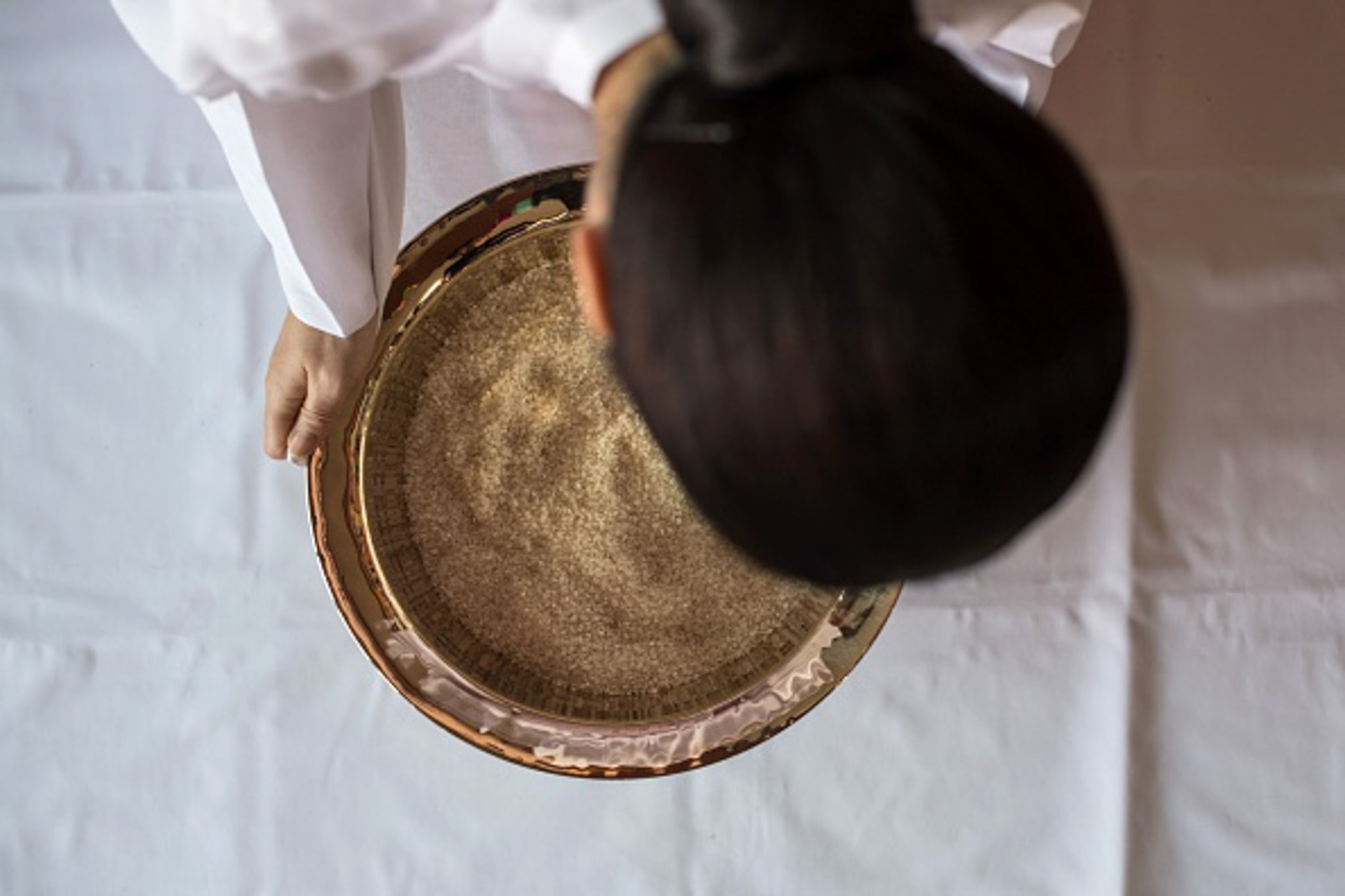 A woman stands over a bowl of rice a white sheet underneath while dressed in white clothing.