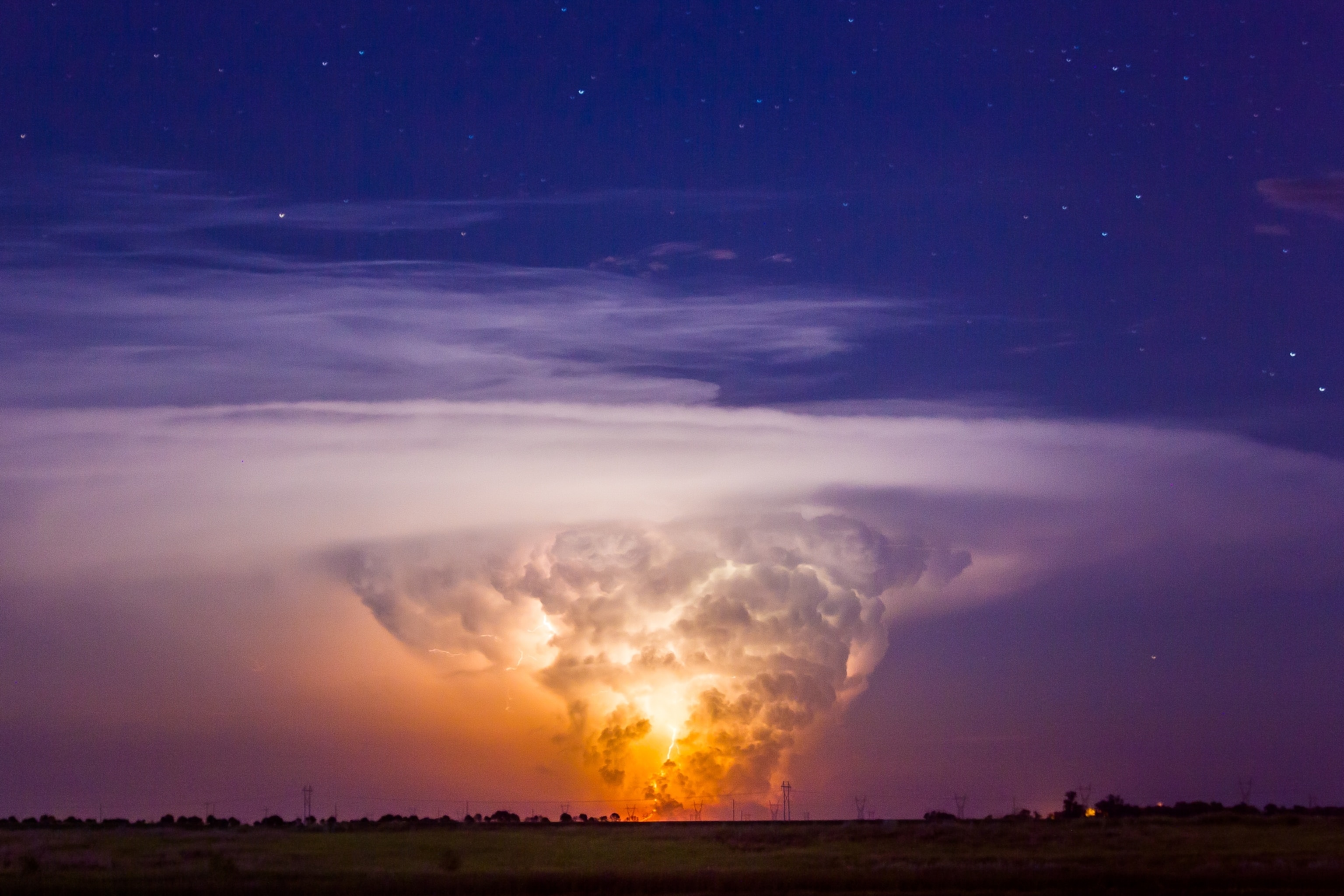 An enormous supercell thunderstorm, with a large billowing tornado touching ground as it is illuminated by lightning within.