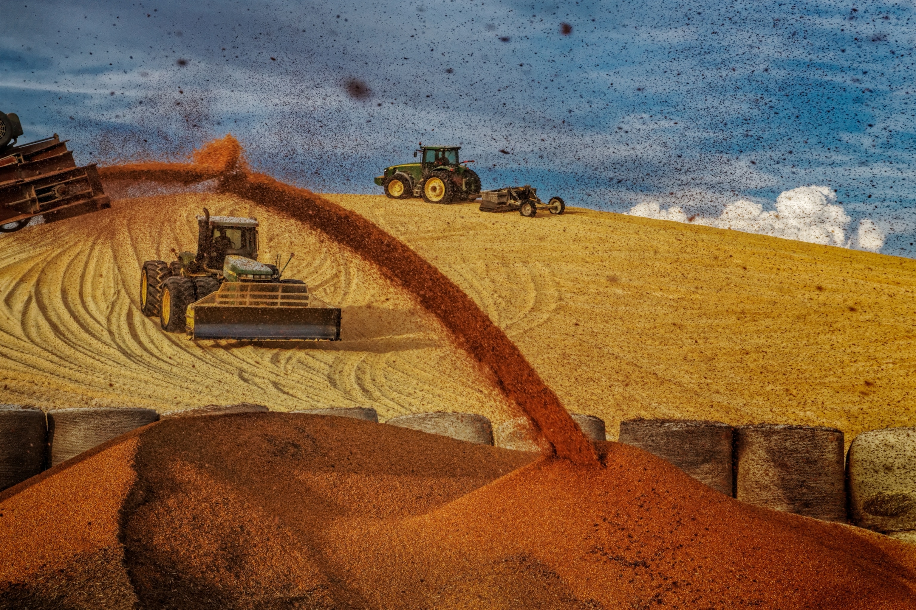 Tractors pack down a giant mound of corn at a feedlot as storm clouds roll in.