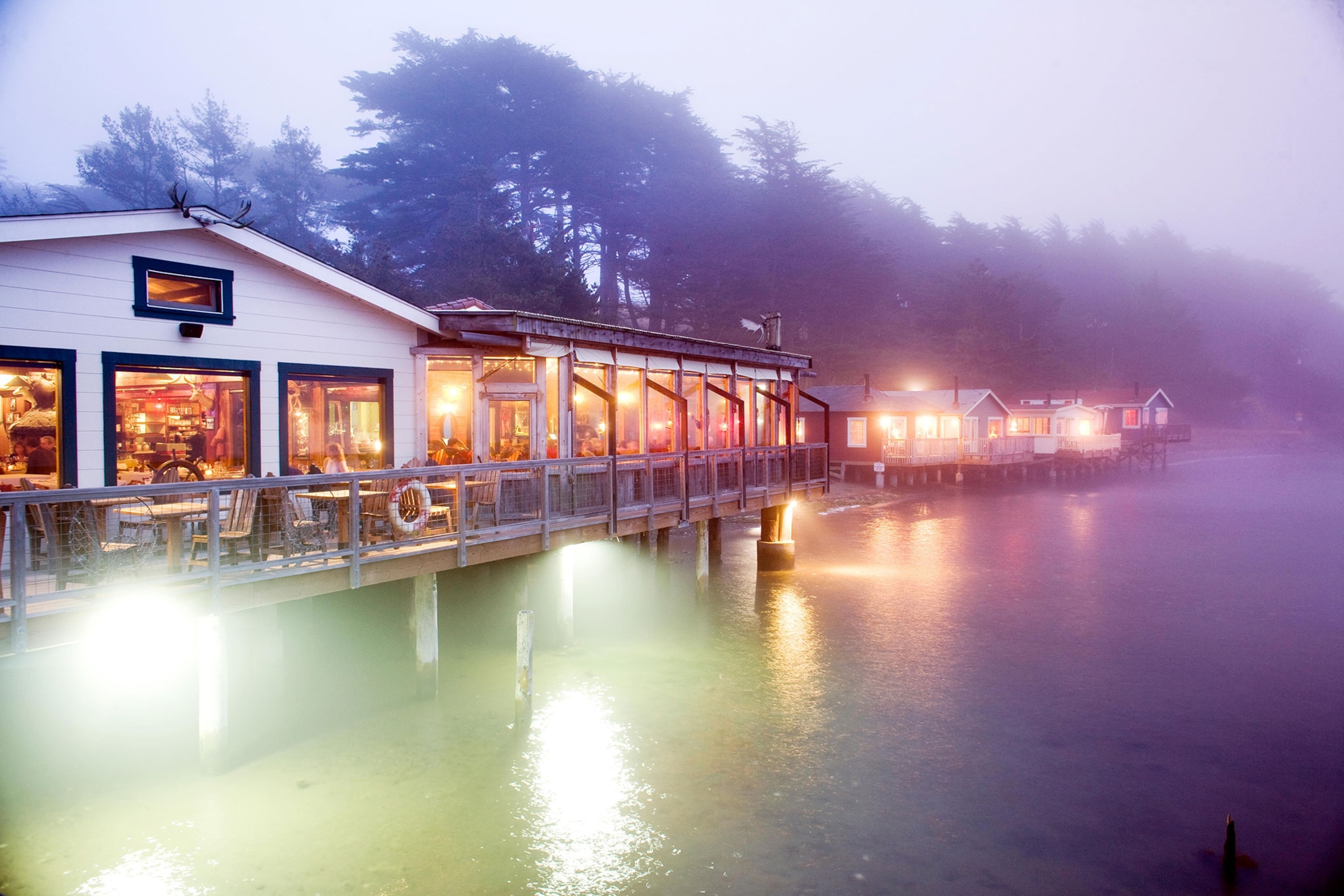 Nick's Cove Restaurant at night over water with fish shacks along the water in the distance.