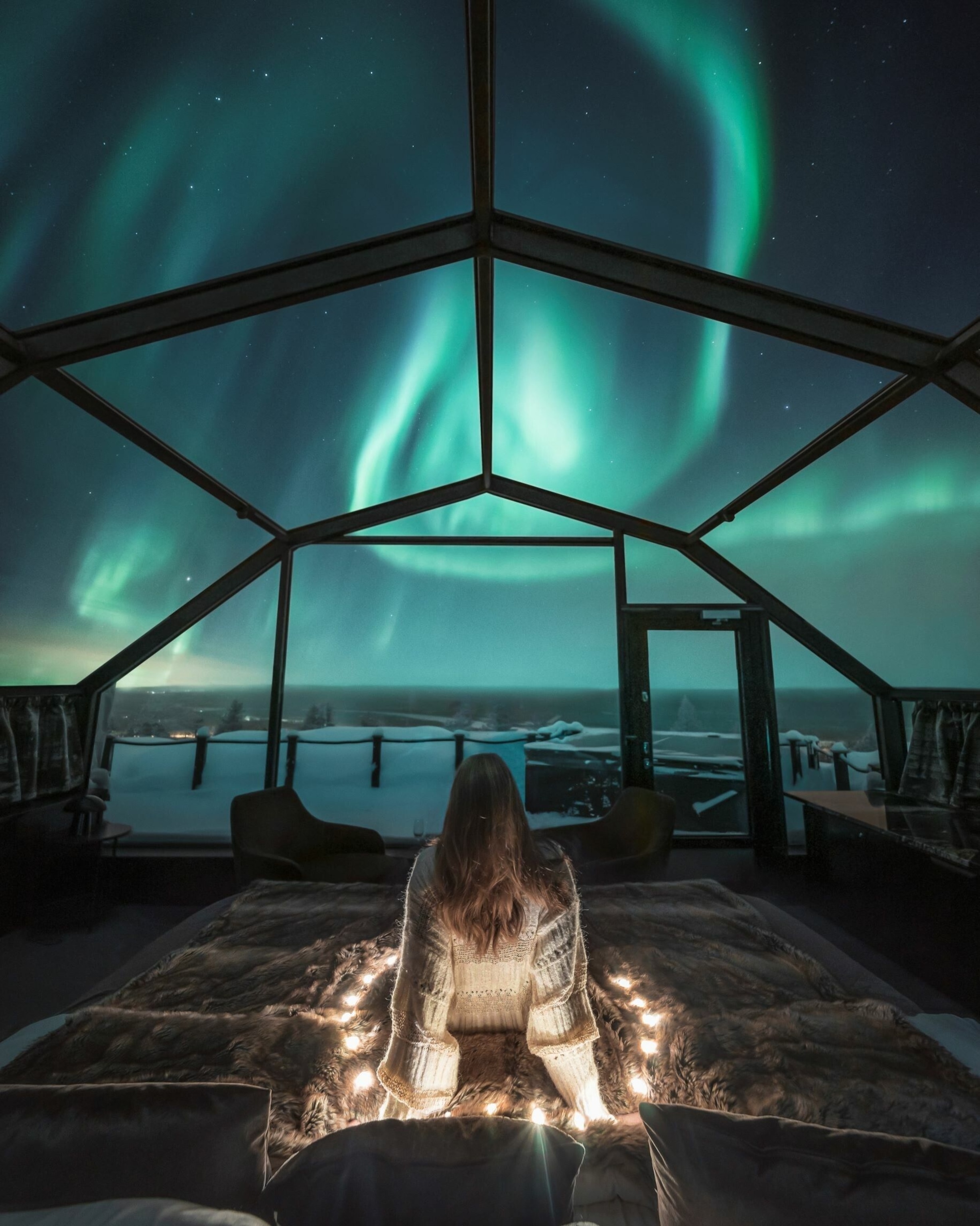 A person on the floor of a glass igloo looking at an aurora.