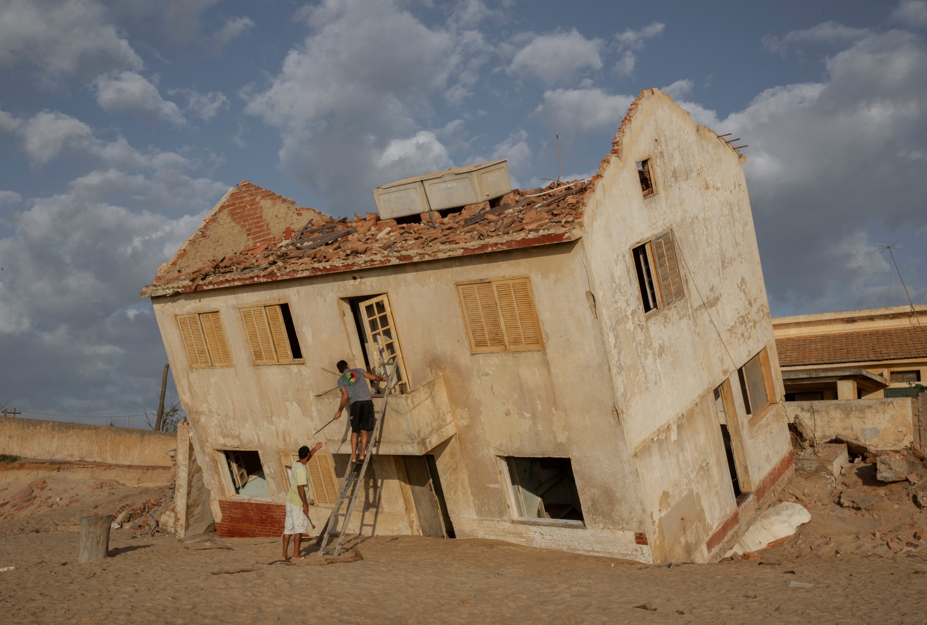 a man on a ladder hands a pipe to a man on the ground as they extract materials from a leaning house
