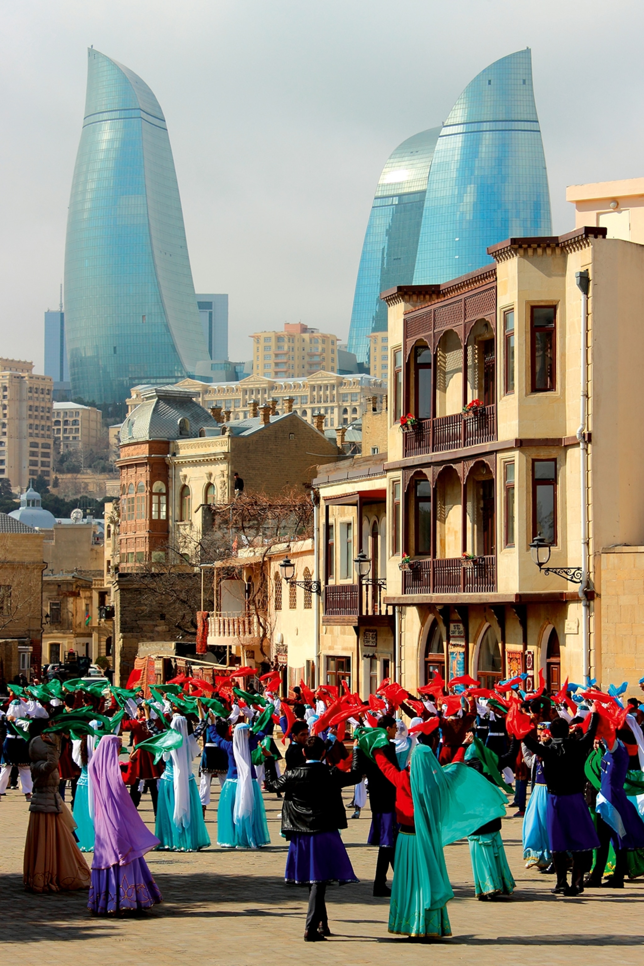 A crowd of pedestrians dressed in colourful clothes, waving flags in a street with sleek glass skyscrapers in the background.