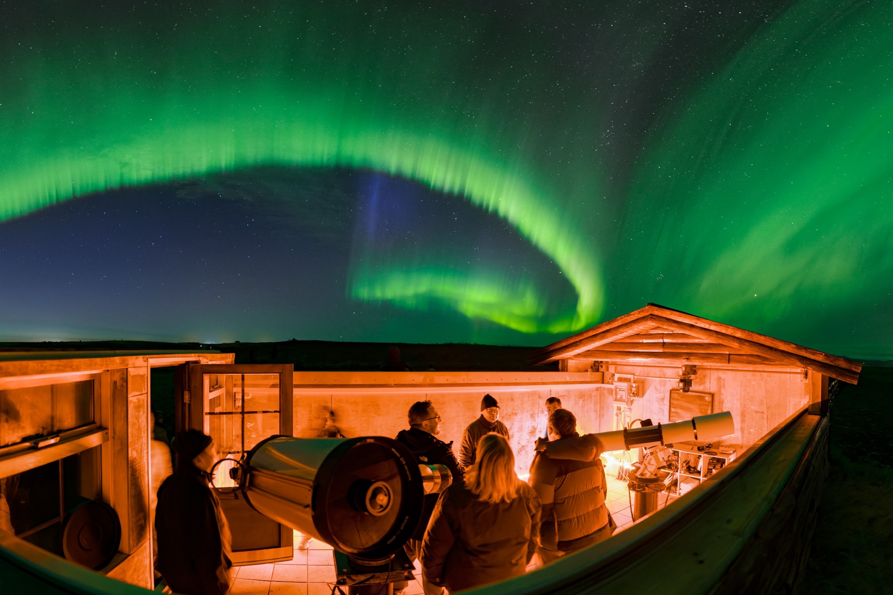 A group of people stand on a roof with telescopes, beneath green ribbons of the Northern Lights streaking across the sky.