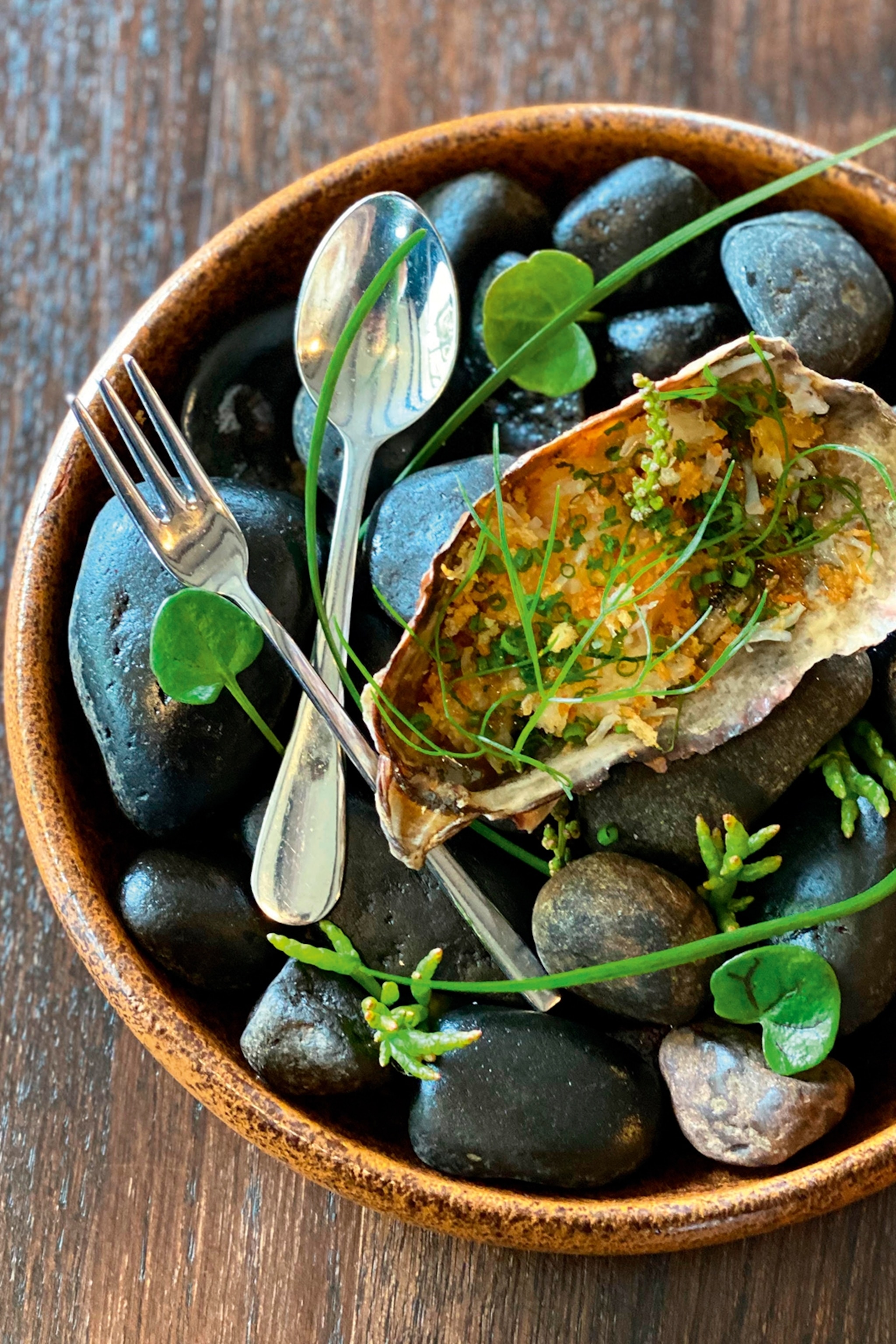Oysters in a bowl garnished with plants presented with a fork and spoon